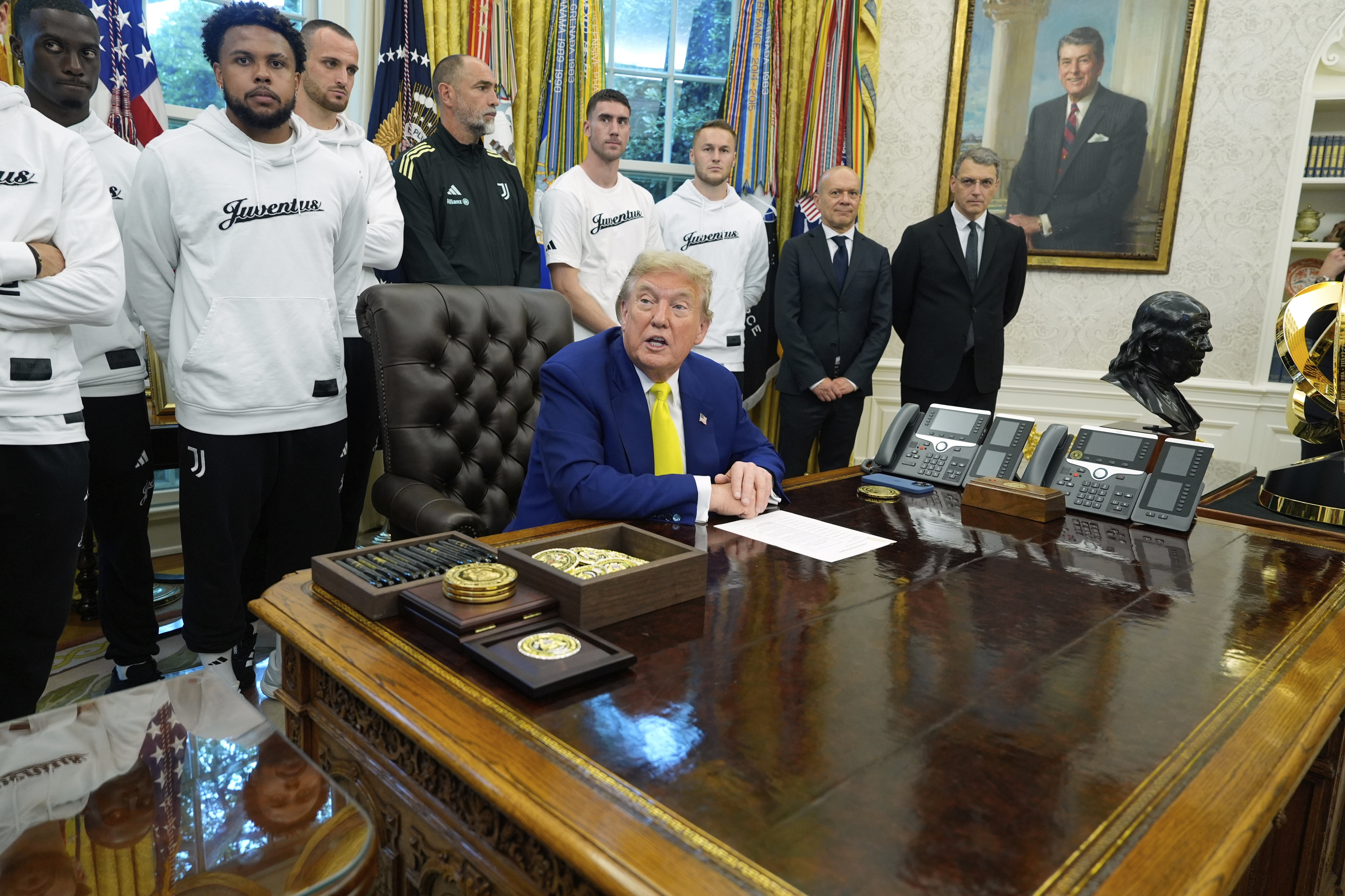 Donald Trump sits at the Resolute Desk, with members of the Juventus soccer team behind him.