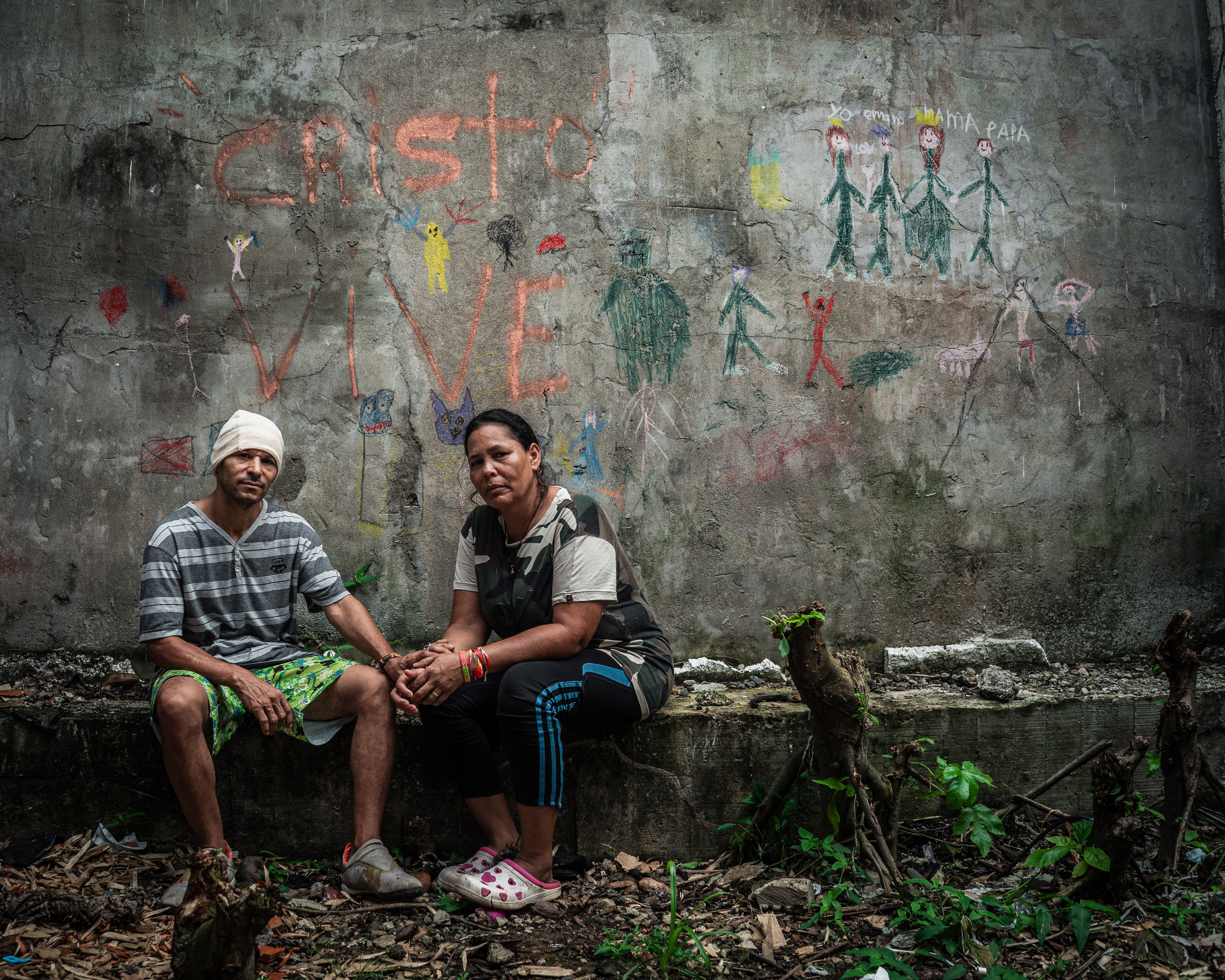 Marisela and Yeral sit on a concrete ledge in front of children's drawings on a bare concrete wall.