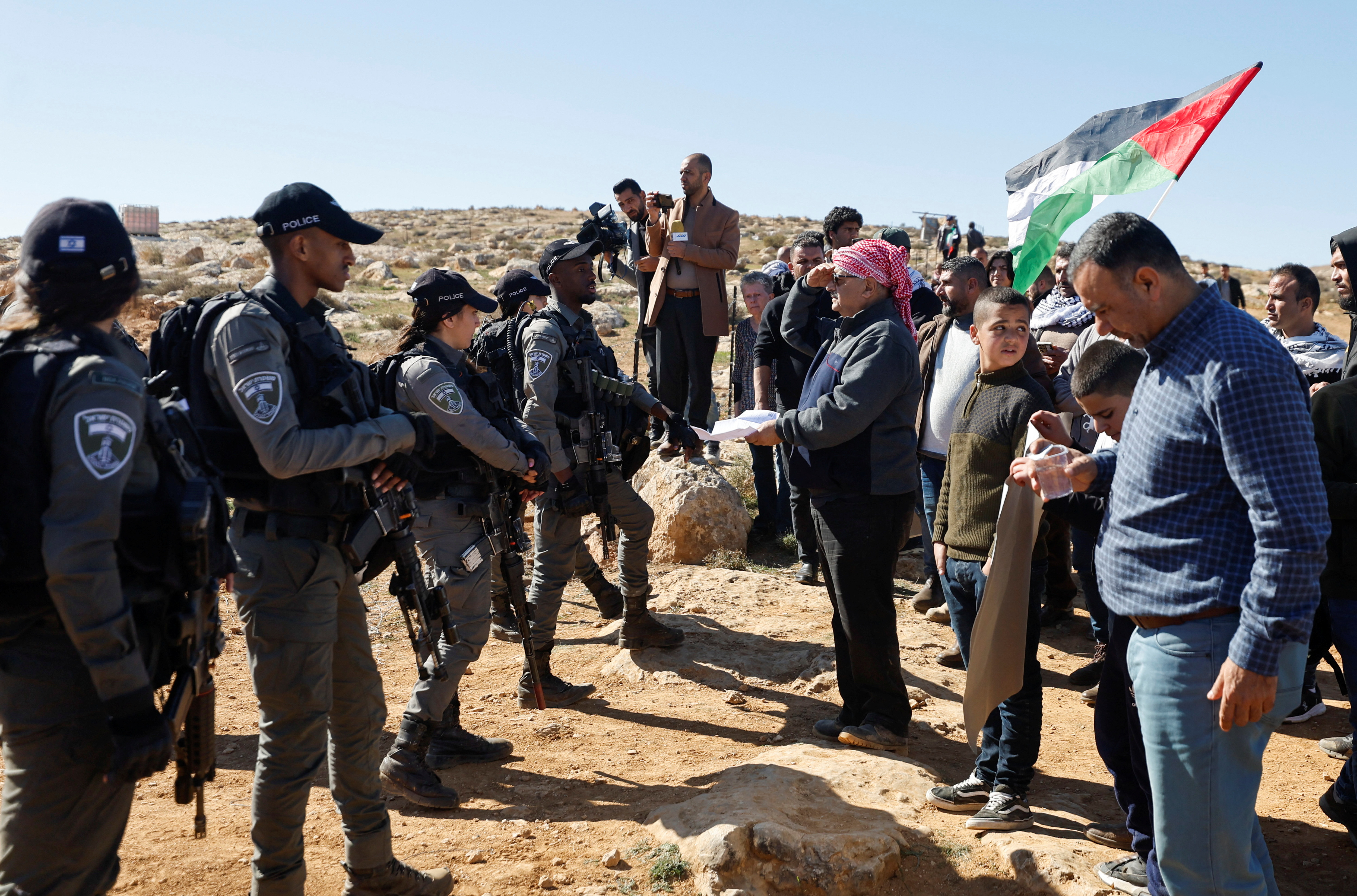 soldiers wearing black stand in a line in front of villagers holding a palestinian flag