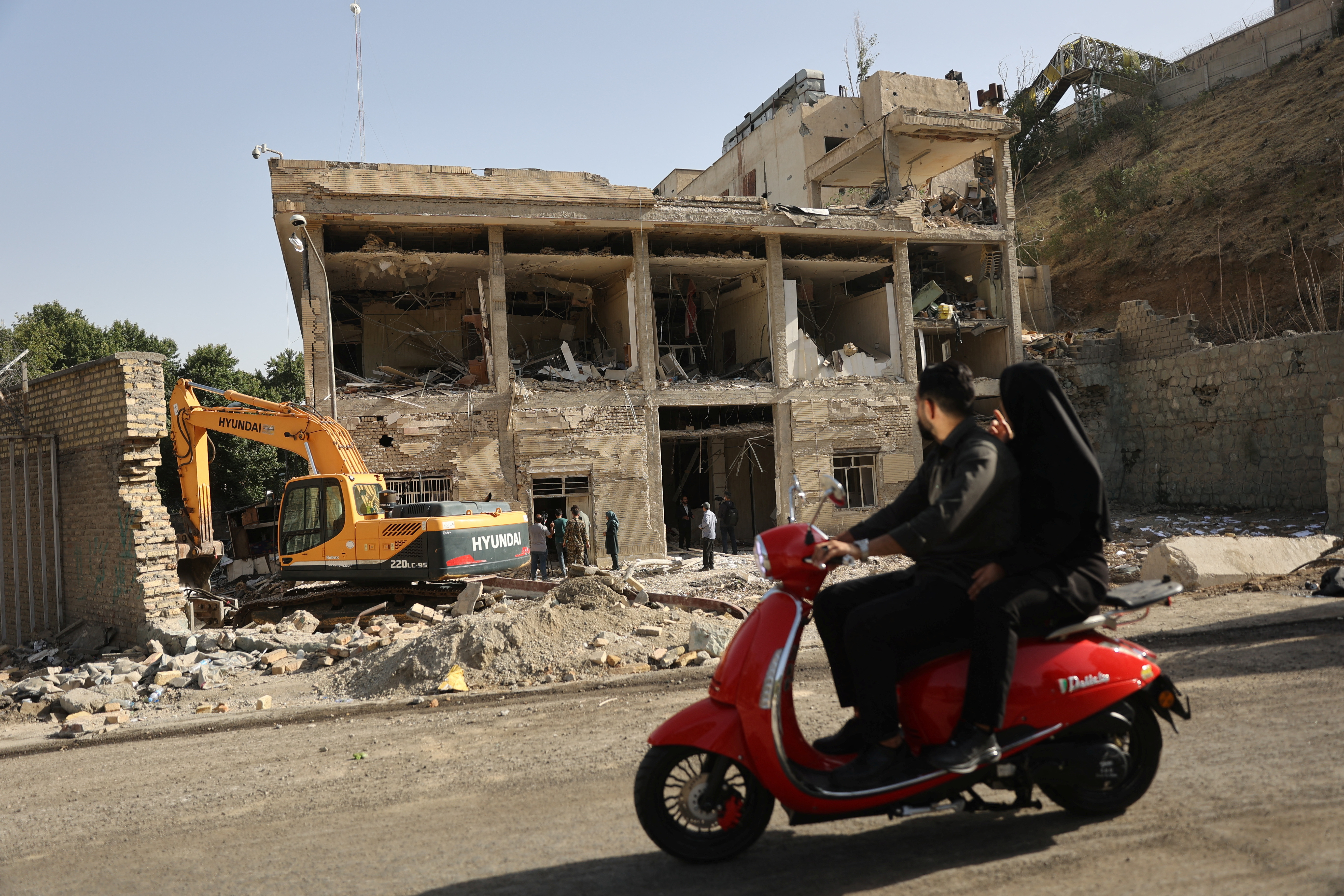 Two people on red motorcycle drives by destroyed building