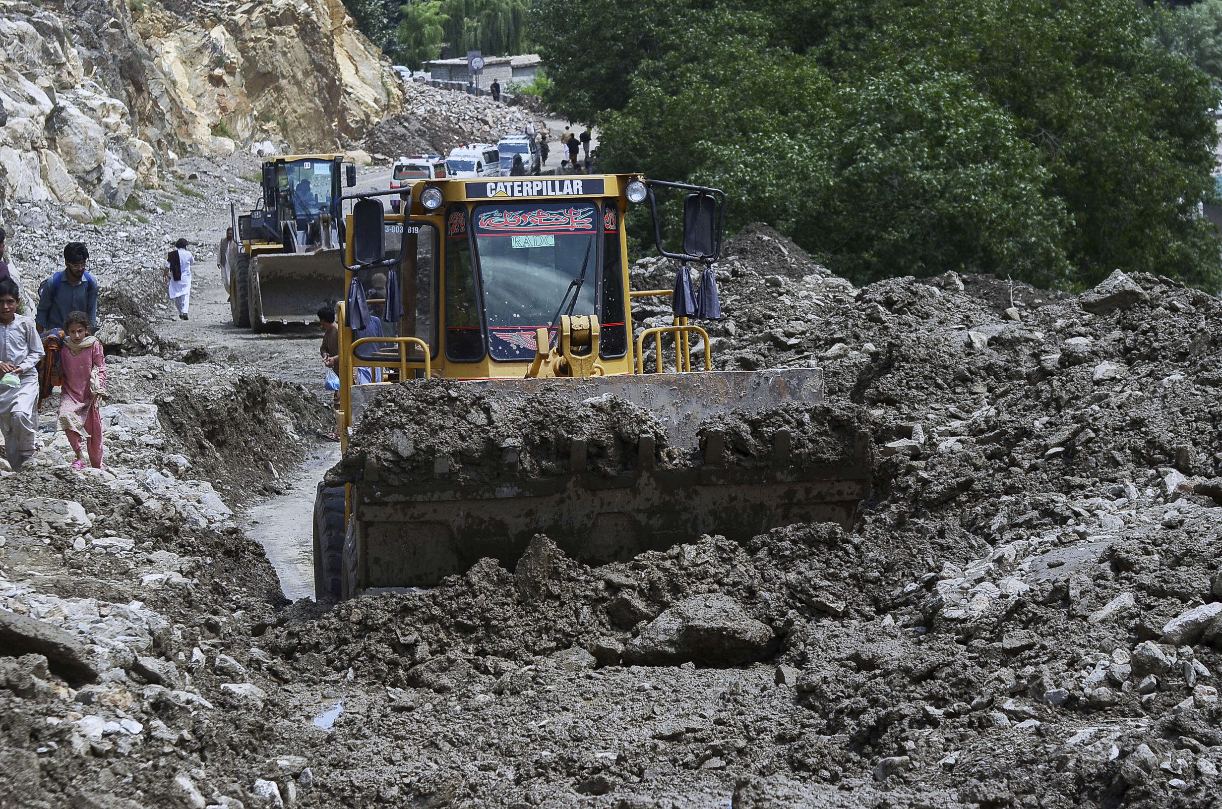 Local residents walk past bulldozers removing debris to clear the way at a site of landslides triggered by flash floods following heavy monsoon rains, on a highway near the Chilas district, northern Pakistan on July 25, 2025.