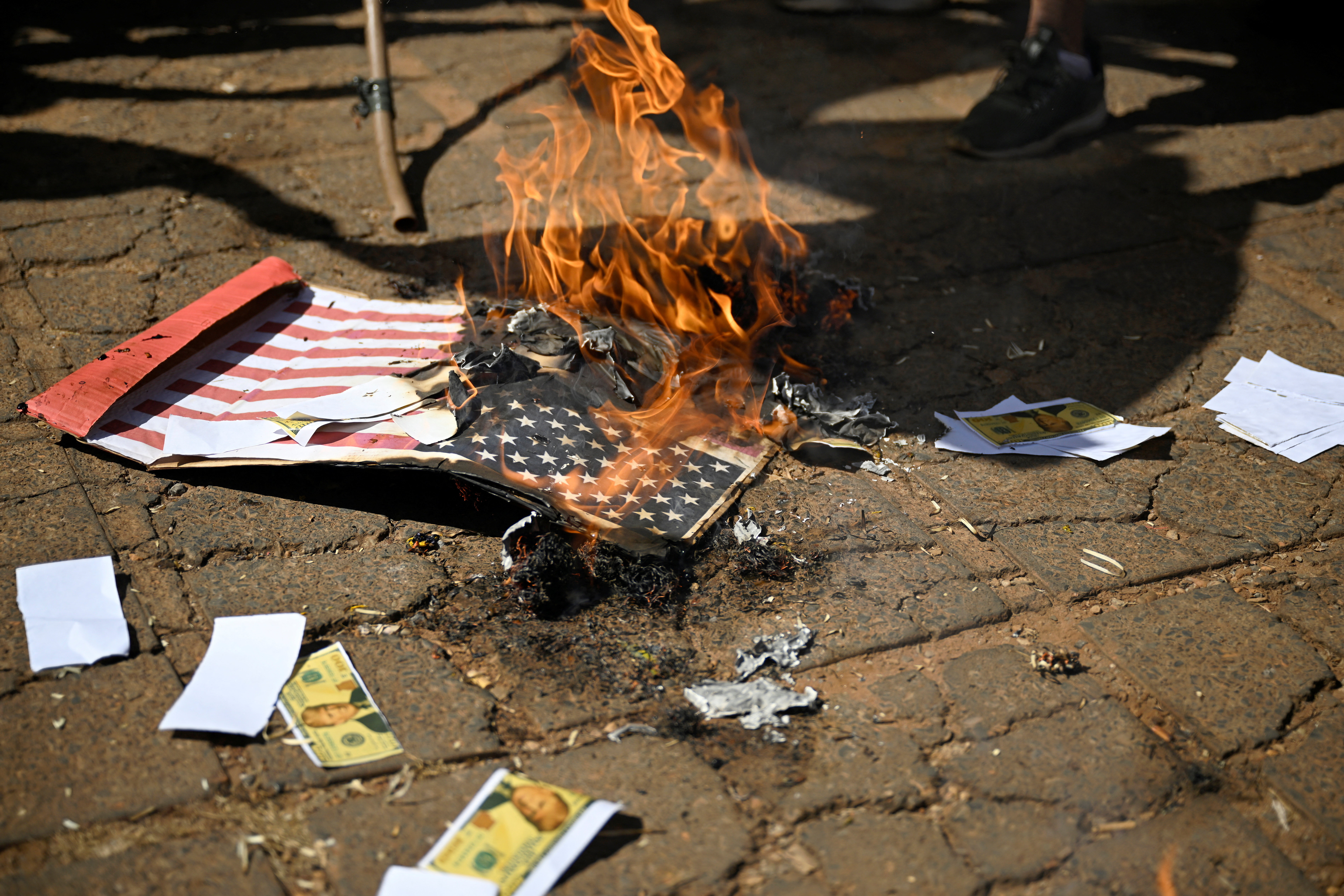 An American flag burns on the ground, next to fake Trump currency.
