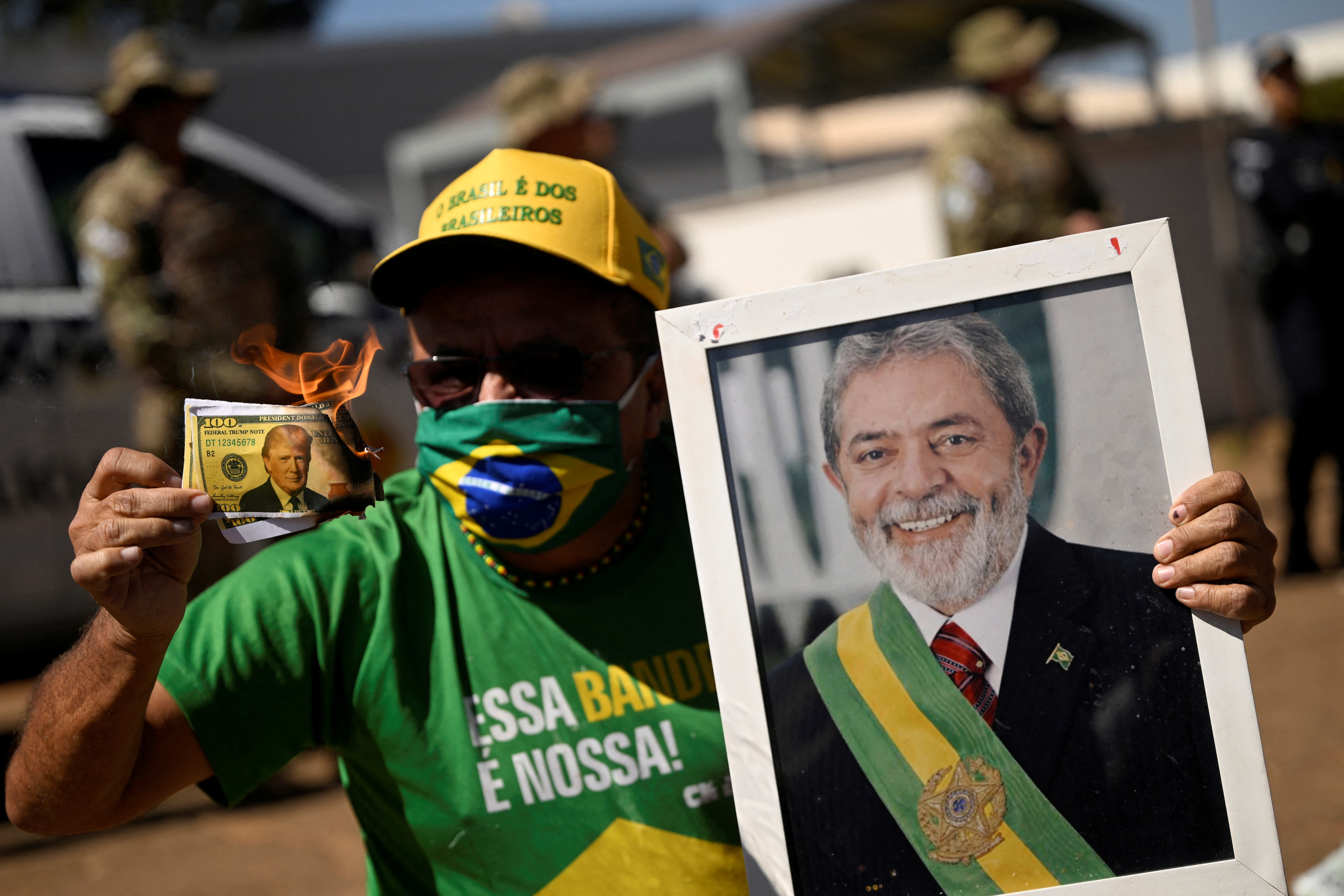 A protester donning a Brazilian flag face mask holds up a portrait of Lula and burning Trump bills.