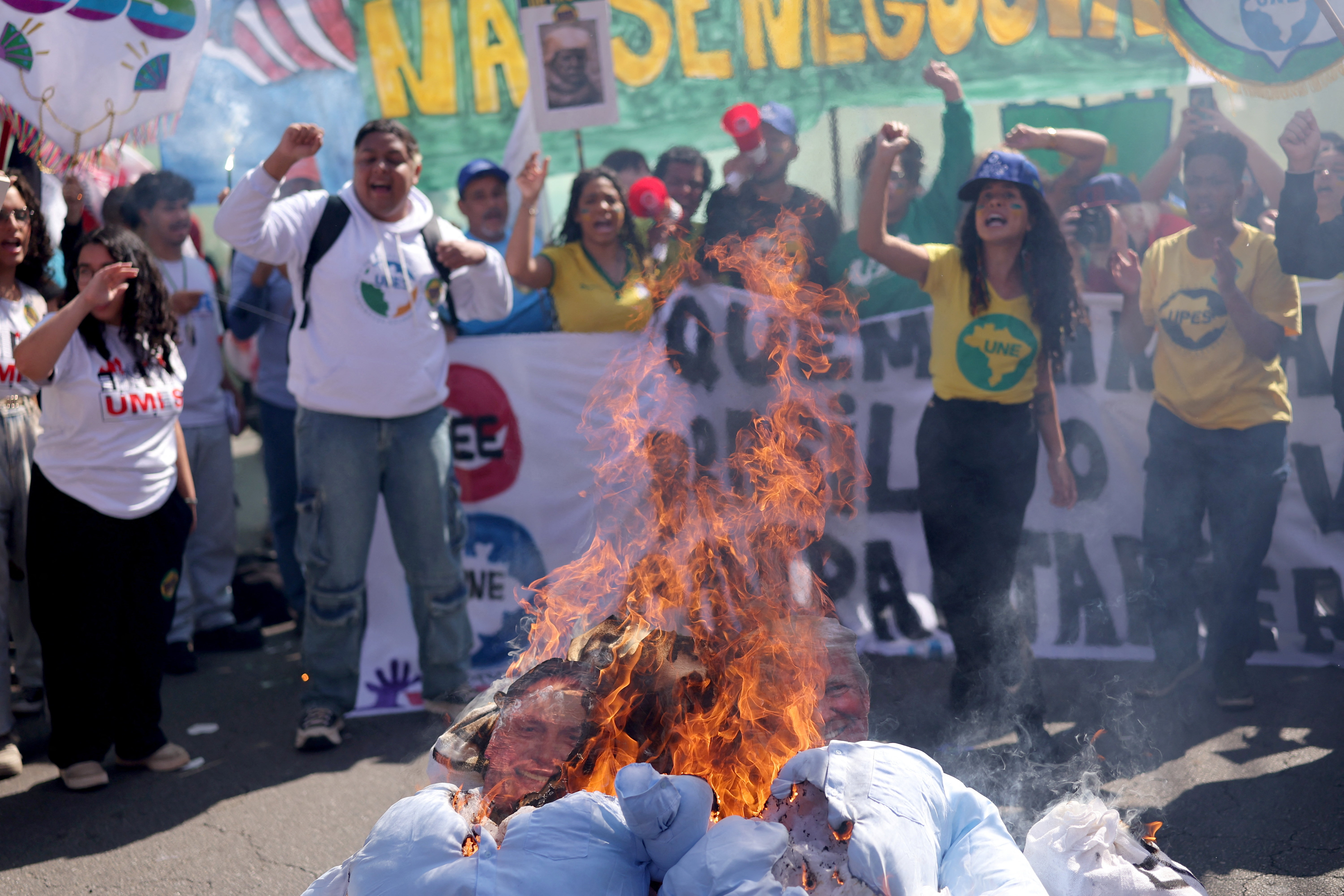 Protesters gather around effigies of Trump and Bolsonaro being burned.