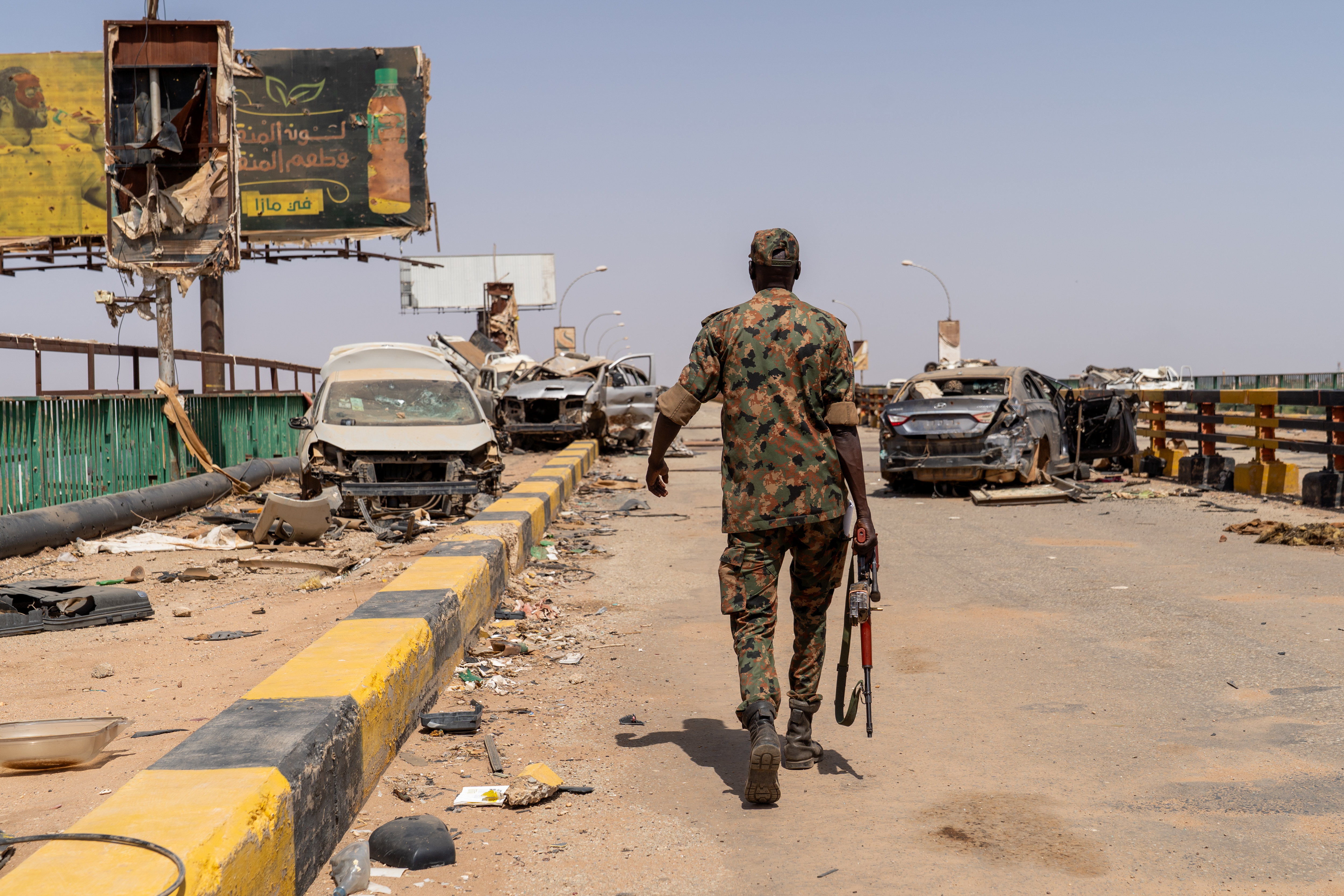 A Sudanese Armed Forces soldier walking on a bridge amidst ruins and destroyed cars.
