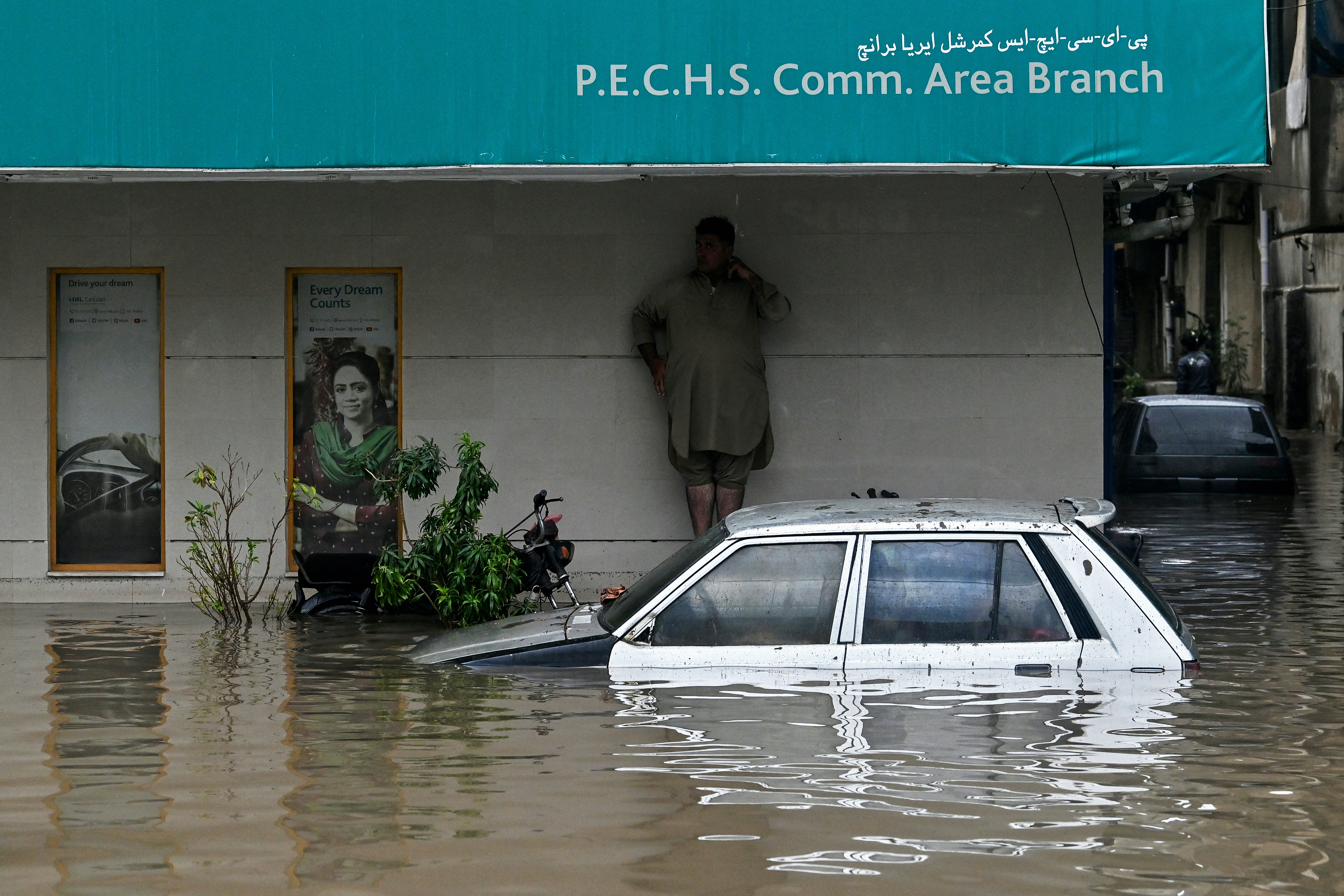 A man takes shelter as a car lies partially submerged in floodwaters along a street after heavy rainfall in Karachi on August 19, 2025.