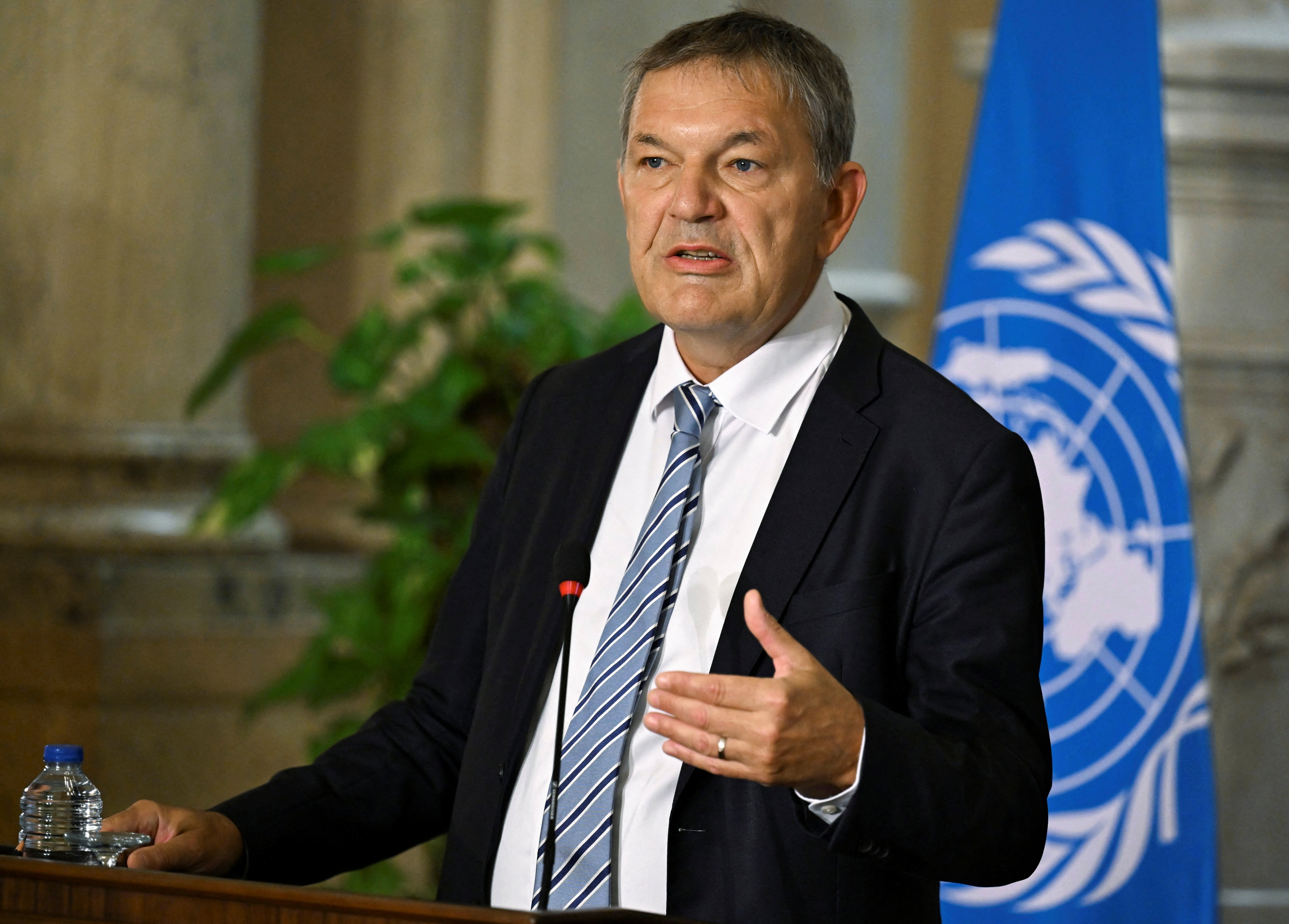 Philippe Lazzarini speaks into a microphone in front of a UN flag