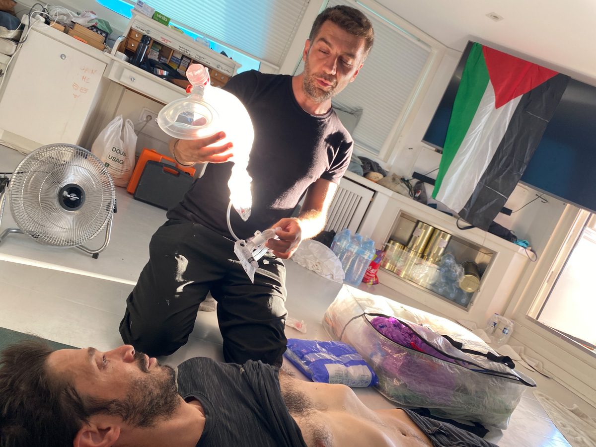 One man lies on the ground, his shirt pulls up, a trainer kneeling beside him is demnonstrating the use of a piece of medical equipment