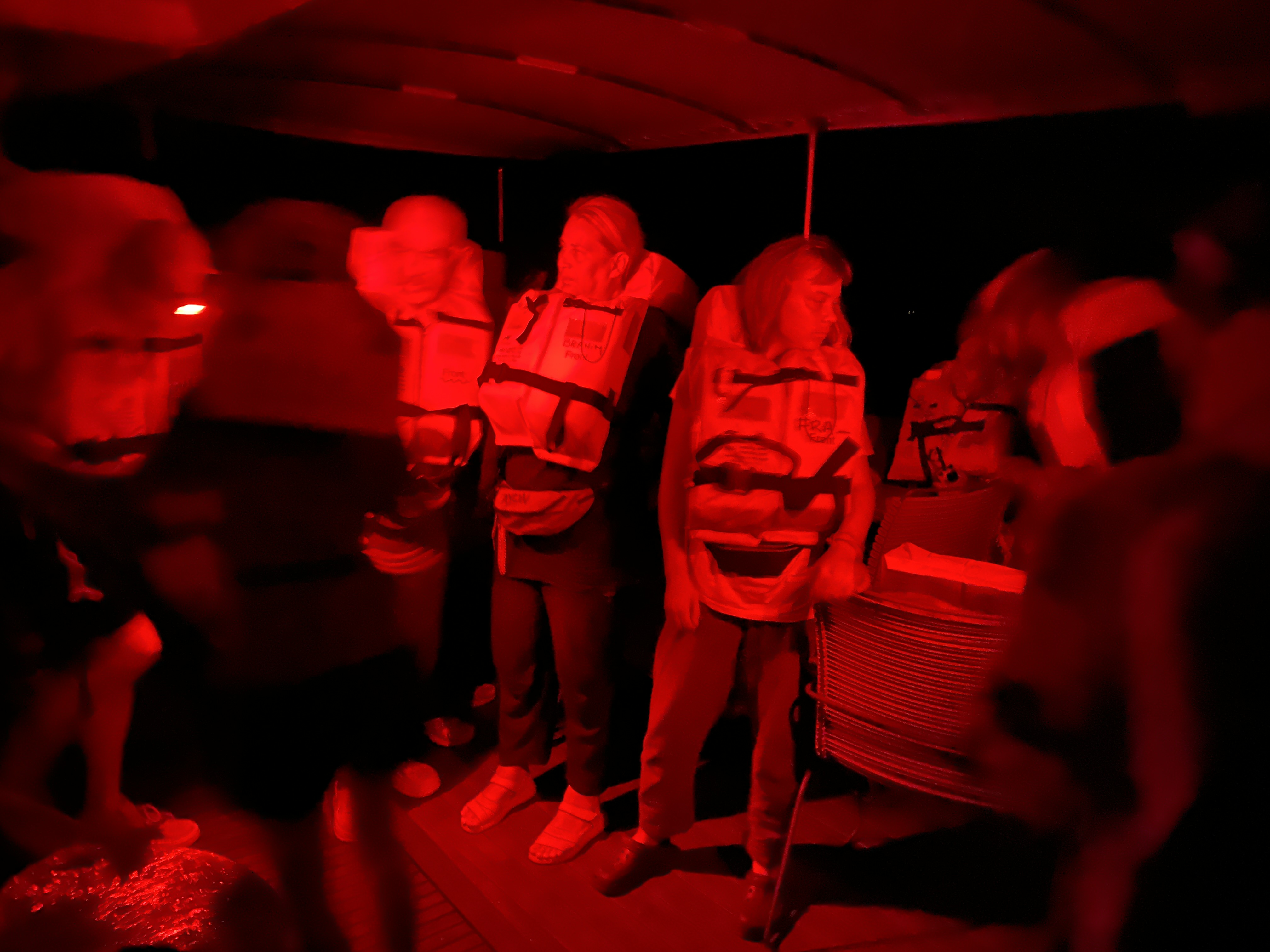 Volunteers appear in red light as it is nighttime, wearing their life vests and listening to a trainer