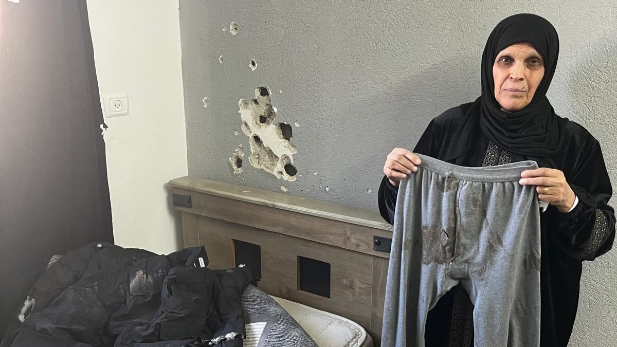 Woman stands holding bloodstained trousers. A bed is to her left, with bulletholes in the wall behind it.