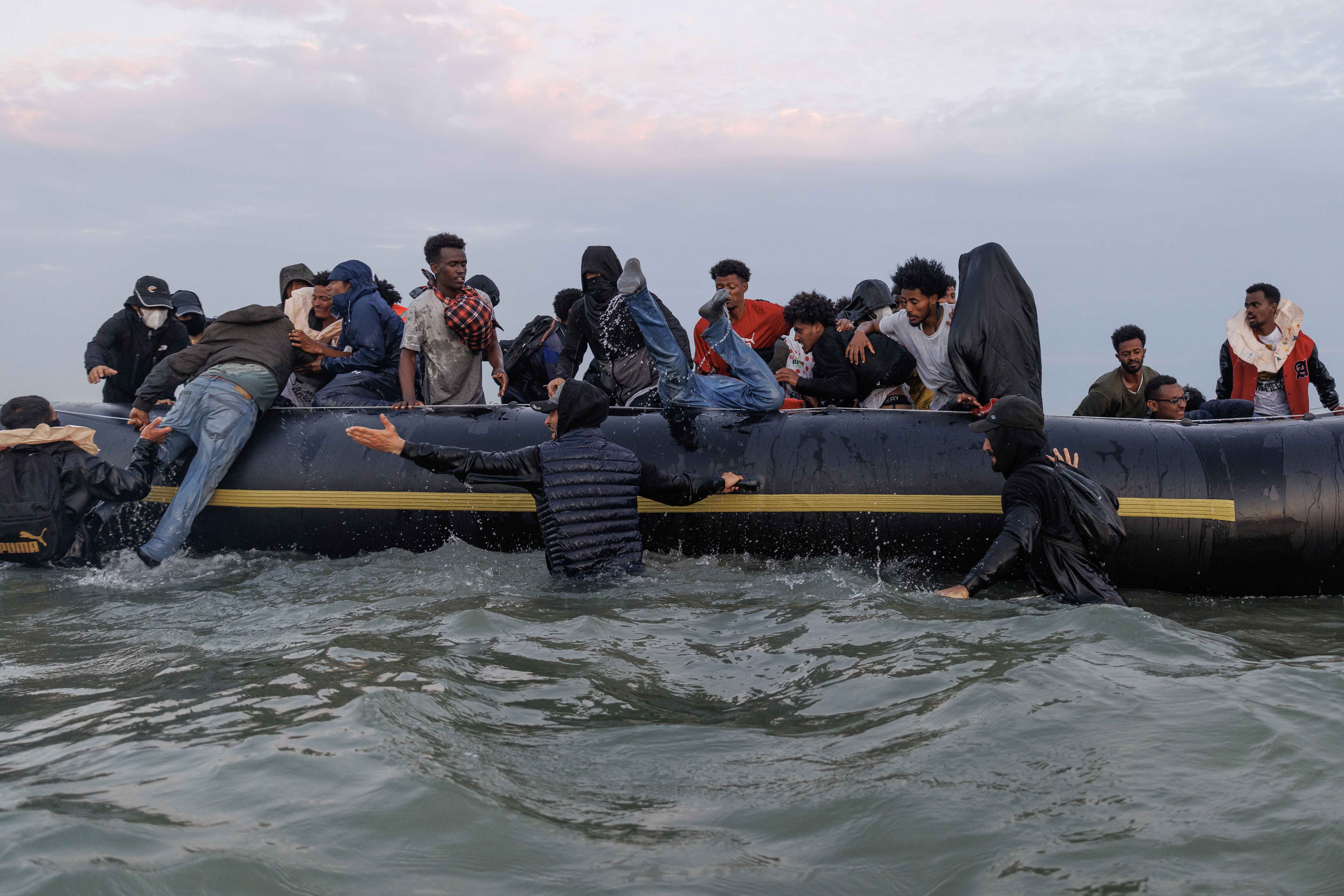 GRAVELINES, FRANCE - JULY 02: Traffickers, (with their faces covered) try to manage the scene using sticks as migrants wade into the water to be collected by a small boat at sunrise on July 02, 2025 in Gravelines, France. Last month, it was reported that French officials were planning changes to policies that govern interceptions at sea, allowing police to intercept small boats within 300 metres of the French shoreline, in a bid to curb migrant crossings of the English Channel. The move, which has not been confirmed, would be welcomed by UK authorities, who see it as a step toward stronger border enforcement. (Photo by Dan Kitwood/Getty Images)