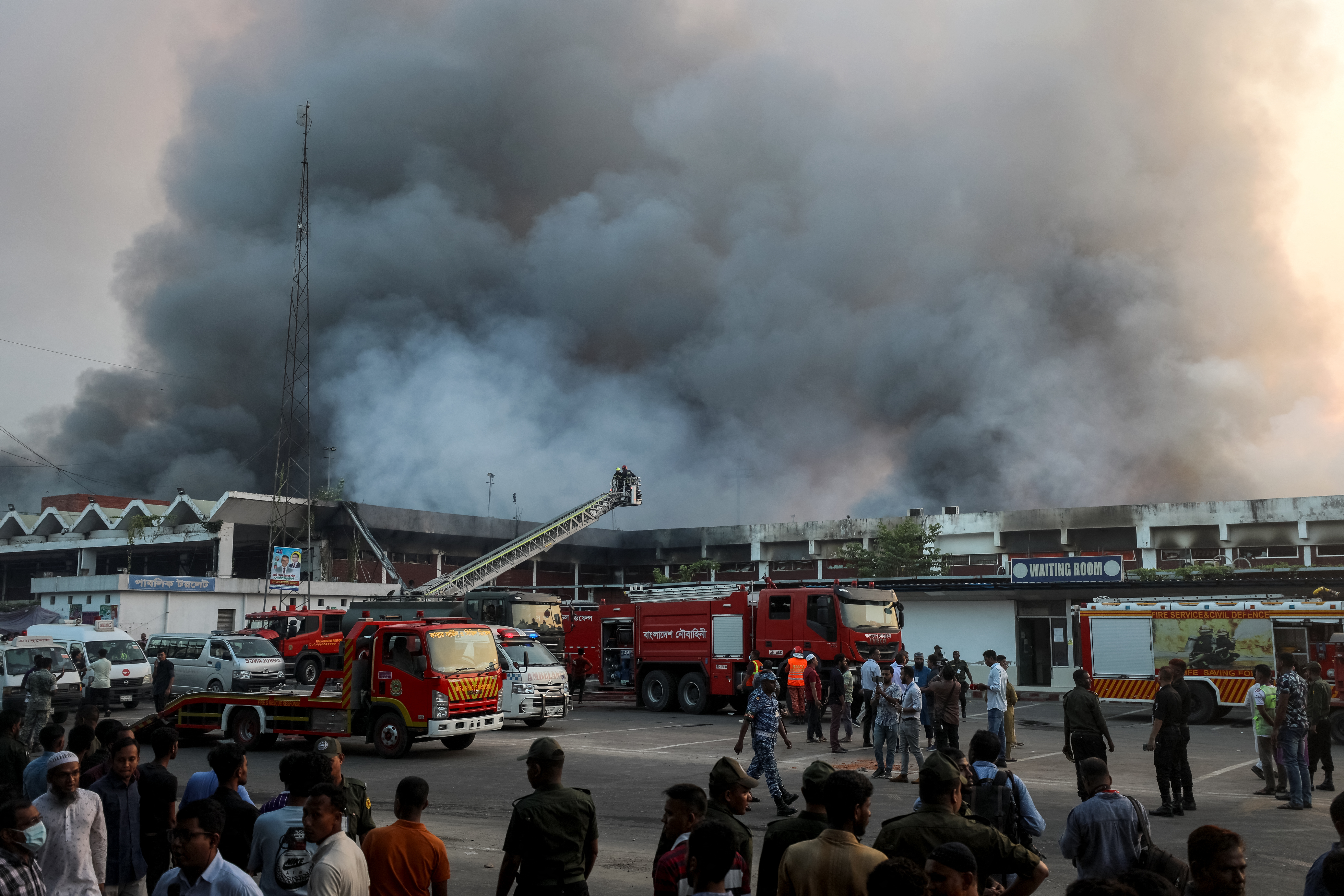 Onlookers gather as firefighters try to extinguish a fire that broke out in the cargo section of Hazrat Shahjalal International Airport in Dhaka on October 18, 2025. A large fire swept through the cargo terminal of Bangladesh's main international airport in Dhaka on October 18, forcing authorities to suspend all flights, officials said. (Photo by Maruf RAHMAN / AFP)