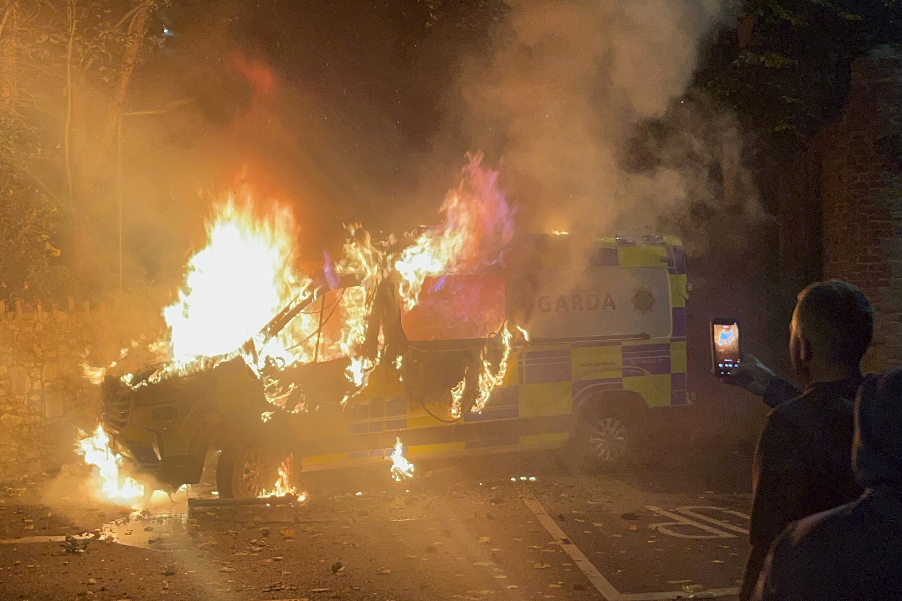 Onlookers film a police vehicle burning as a demonstration outside a hotel housing asylum seekers turns violent [Peter Murphy/AFP]