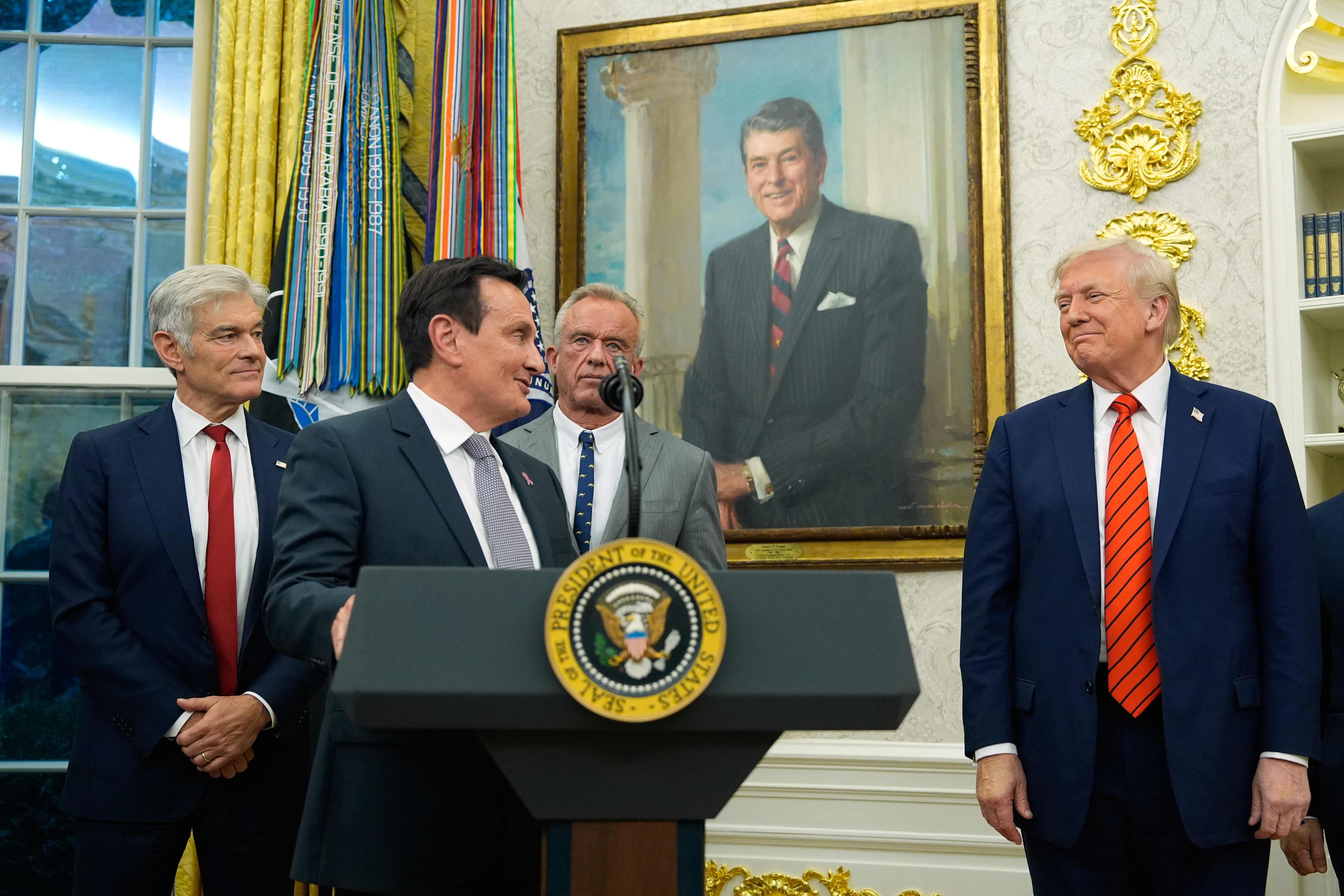 Pascal Soriot speaks behind a presidential podium in the Oval Office, standing next to Trump.