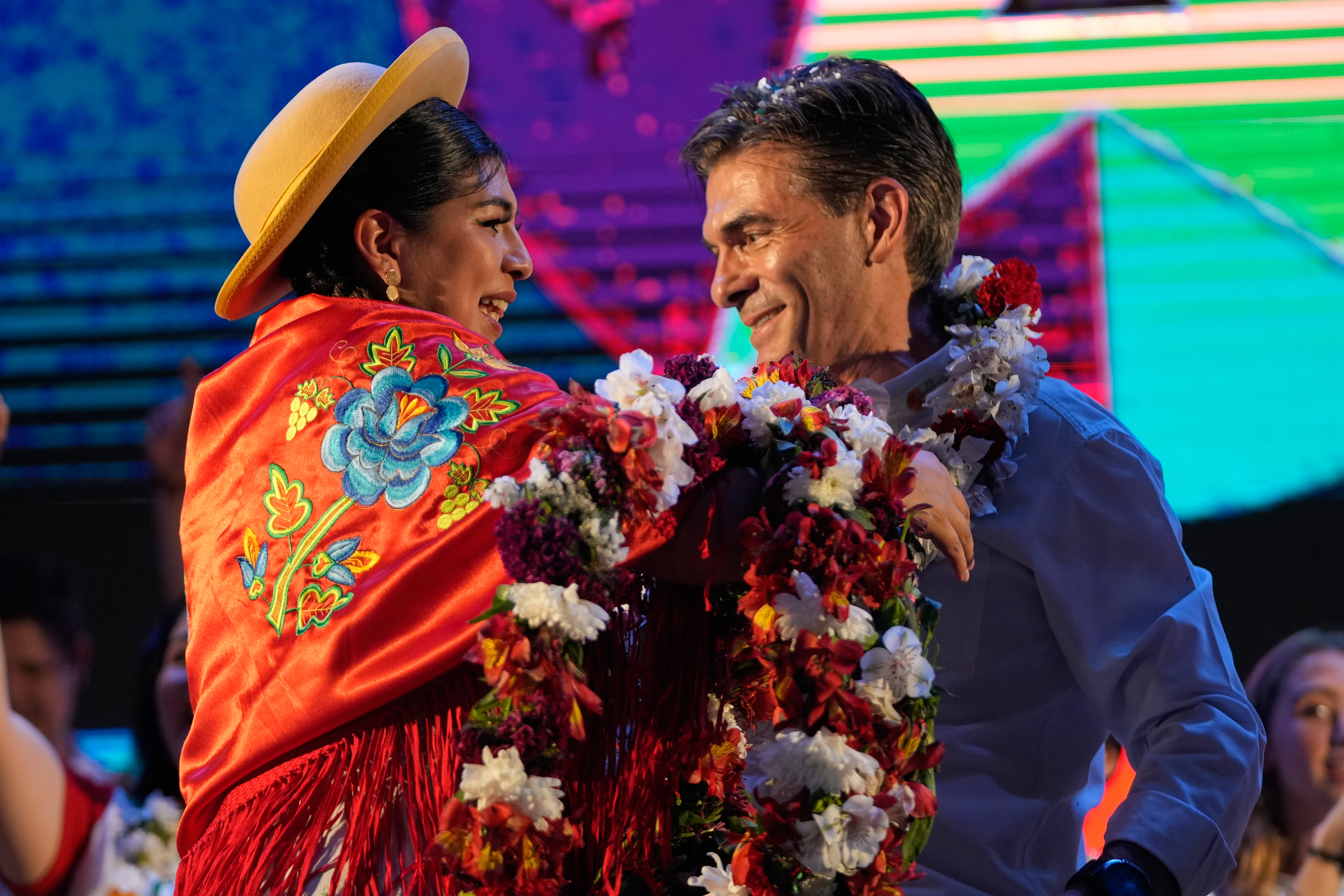 A Bolivian woman places a garland of flowers around Rodrigo Paz's neck on stage at a campaign event.