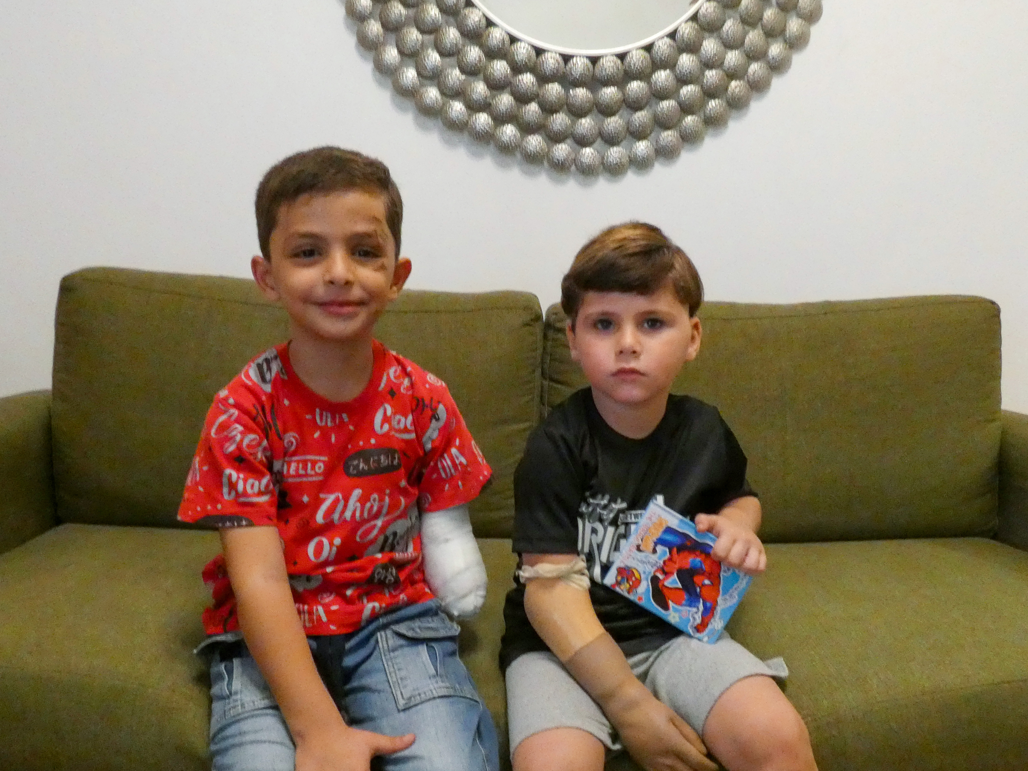 Omar on the left is smiling slightly at the camera, his stump hanging by his left side. Ali stares more seriously at the camera, holding on to his Spider-Man notebook. They are on an olive-green sofa in a living room