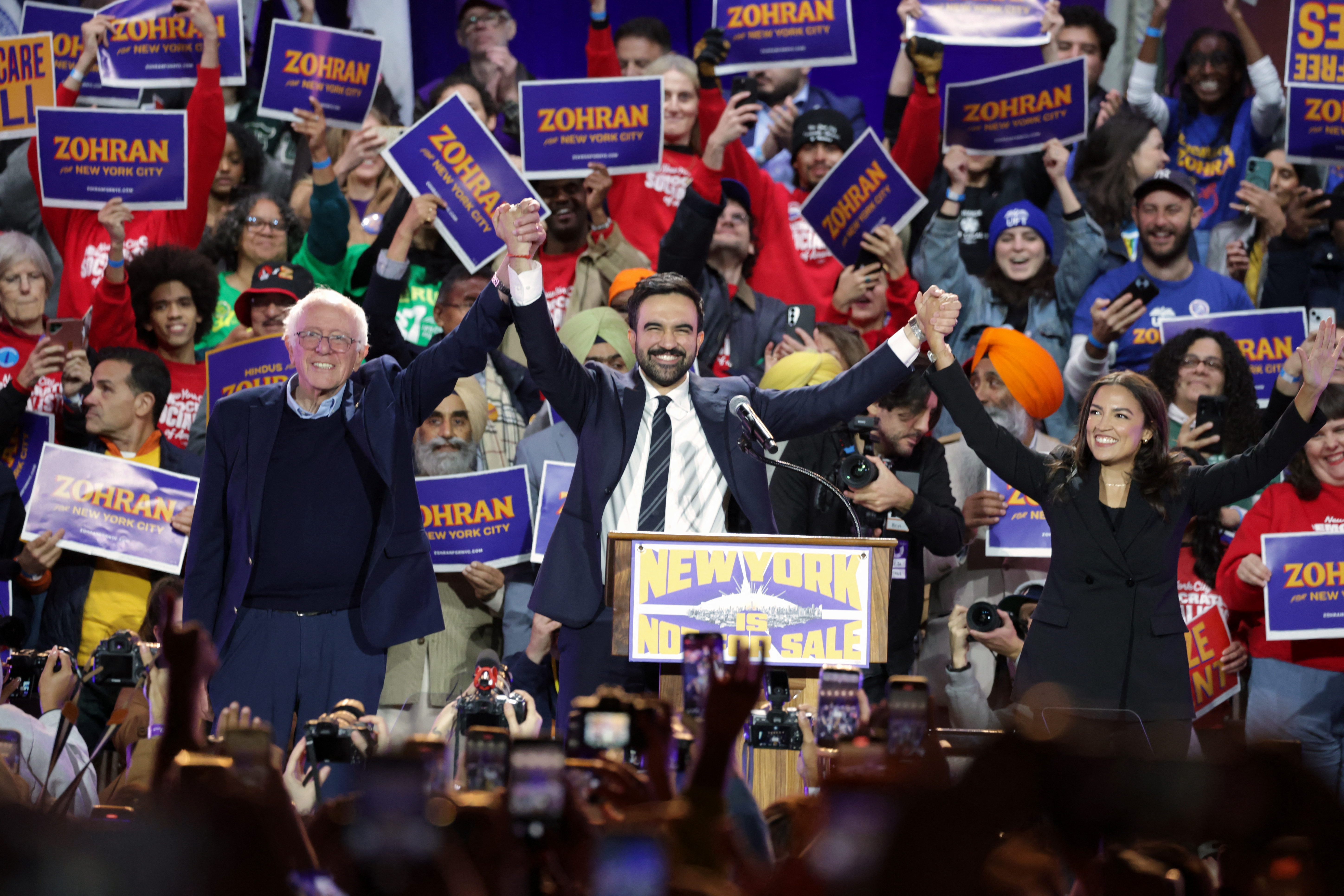 New York City mayoral candidate Zohran Mamdani (C) holds hands with US Senator Bernie Sanders (I-VT) (L) and US Representative Alexandria Ocasio-Cortez (D-NY) at the end of a campaign rally at Forest Hills Stadium in the Queens borough of New York City on October 26, 2025.