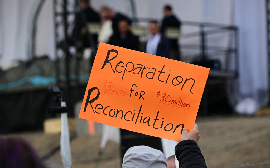 People hold protest sign demanding monetary reparation for reconciliation, as the late Pope Francis arrives to meet with Indigenous people at Nakasuk school in Iqaluit, Nunavut, Canada, on July 29, 2022
