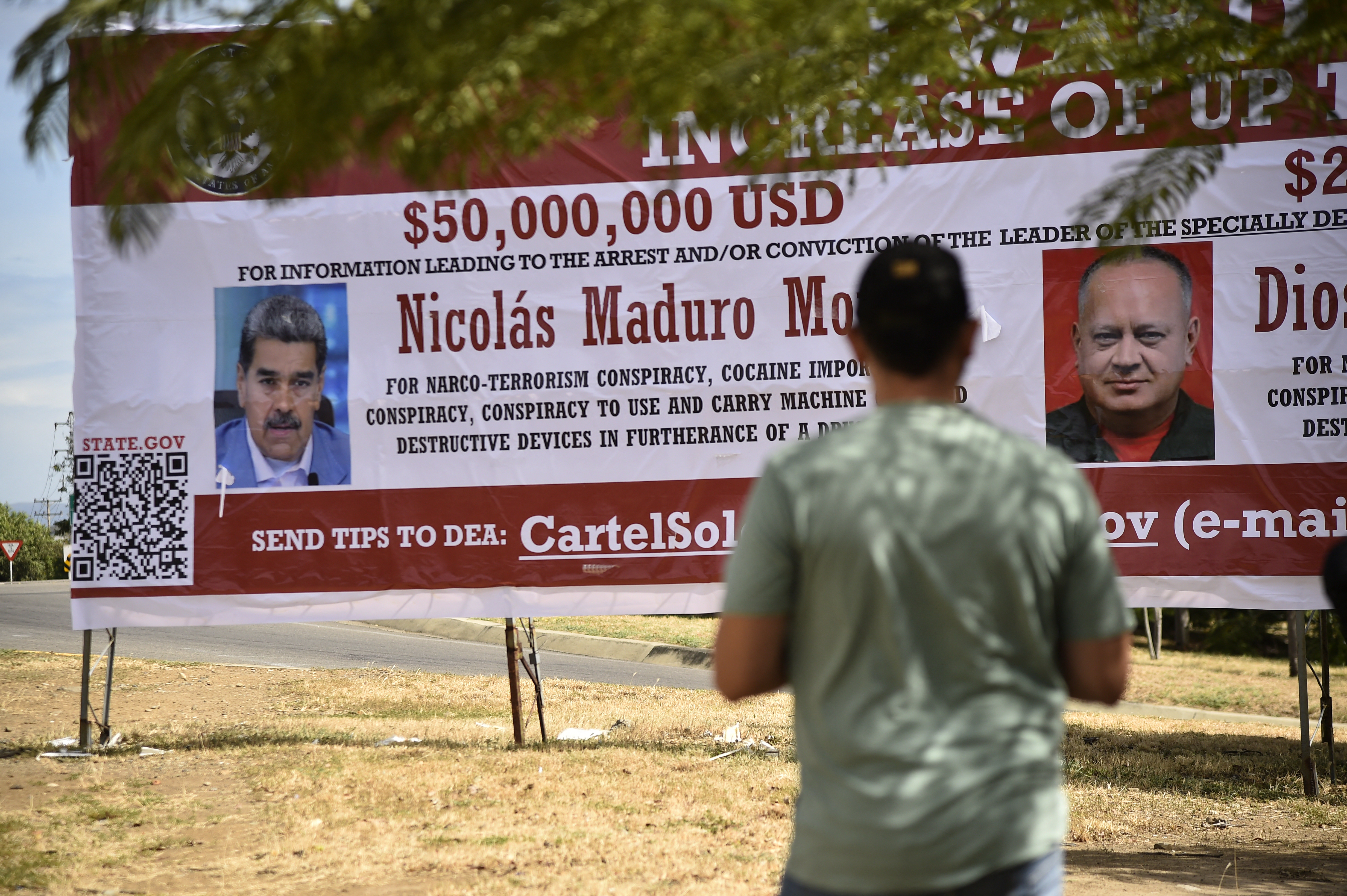 A man stands in front of a billboard advertising the reward for Maduro