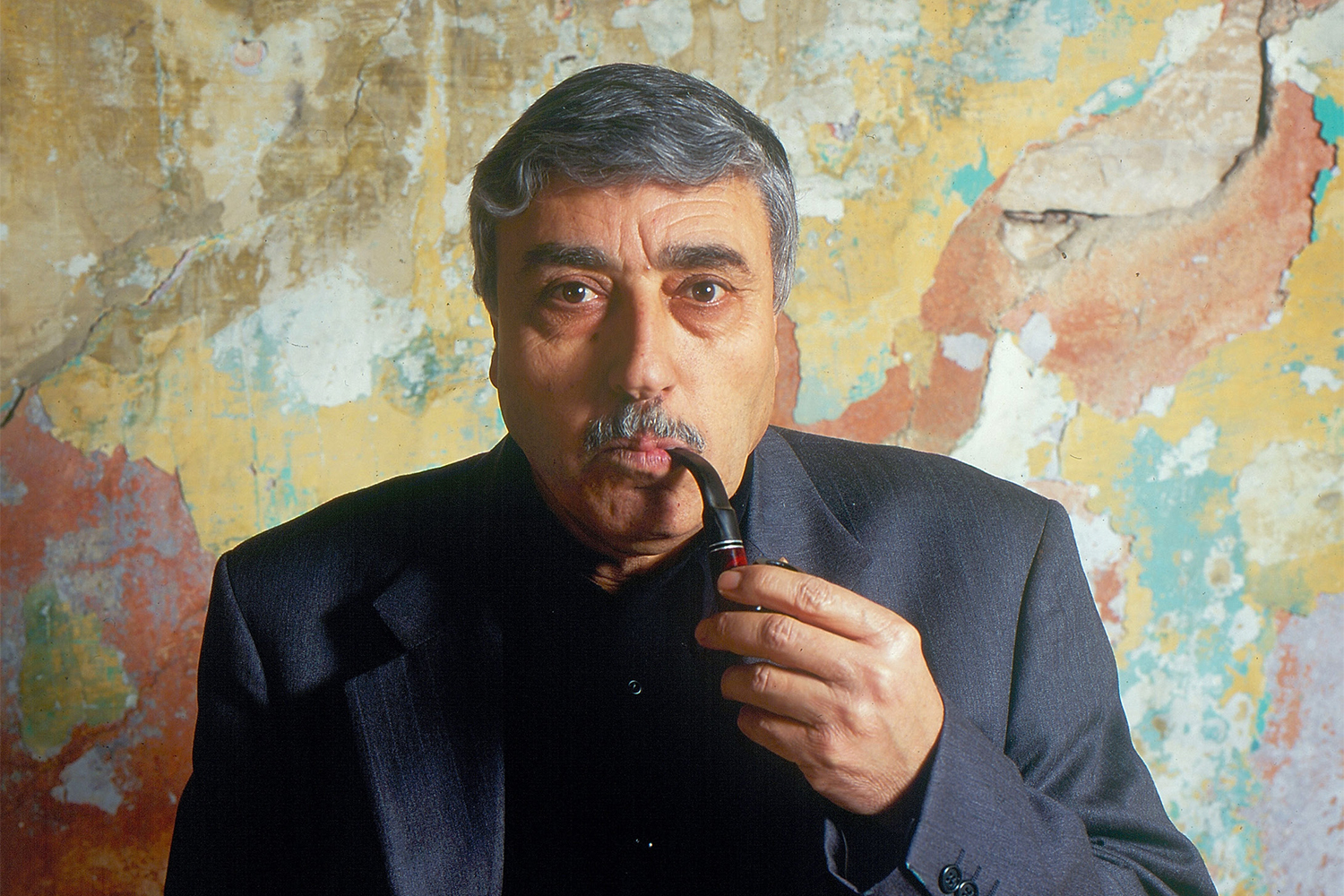Nabil looks right at the camera, a pipe in his mouth, held in his left hand. Behind him is a large artwork in earth tones