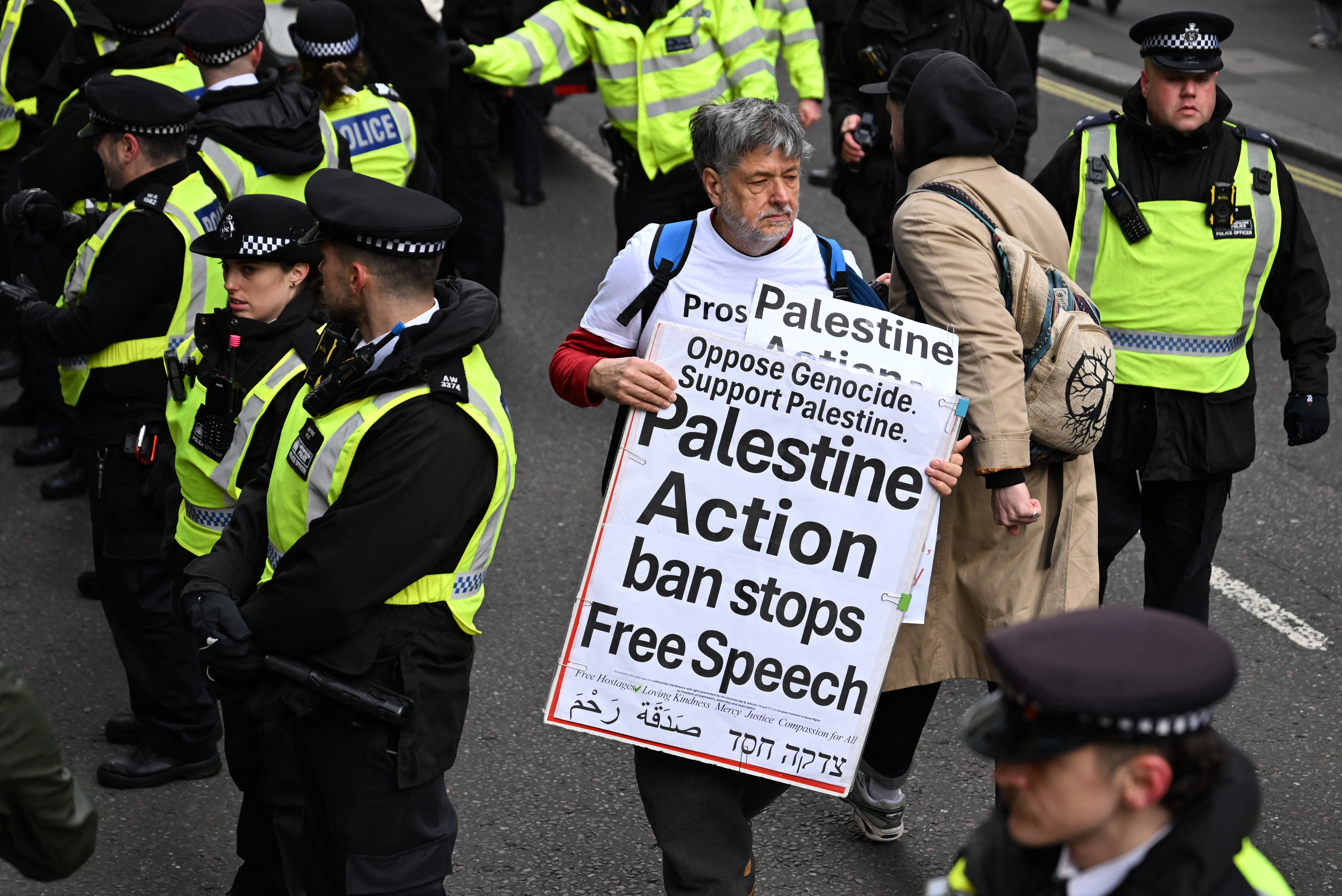 A protester holds placard reading 'Palestine Action ban stops Free Speech' whilst surrounded by police officers