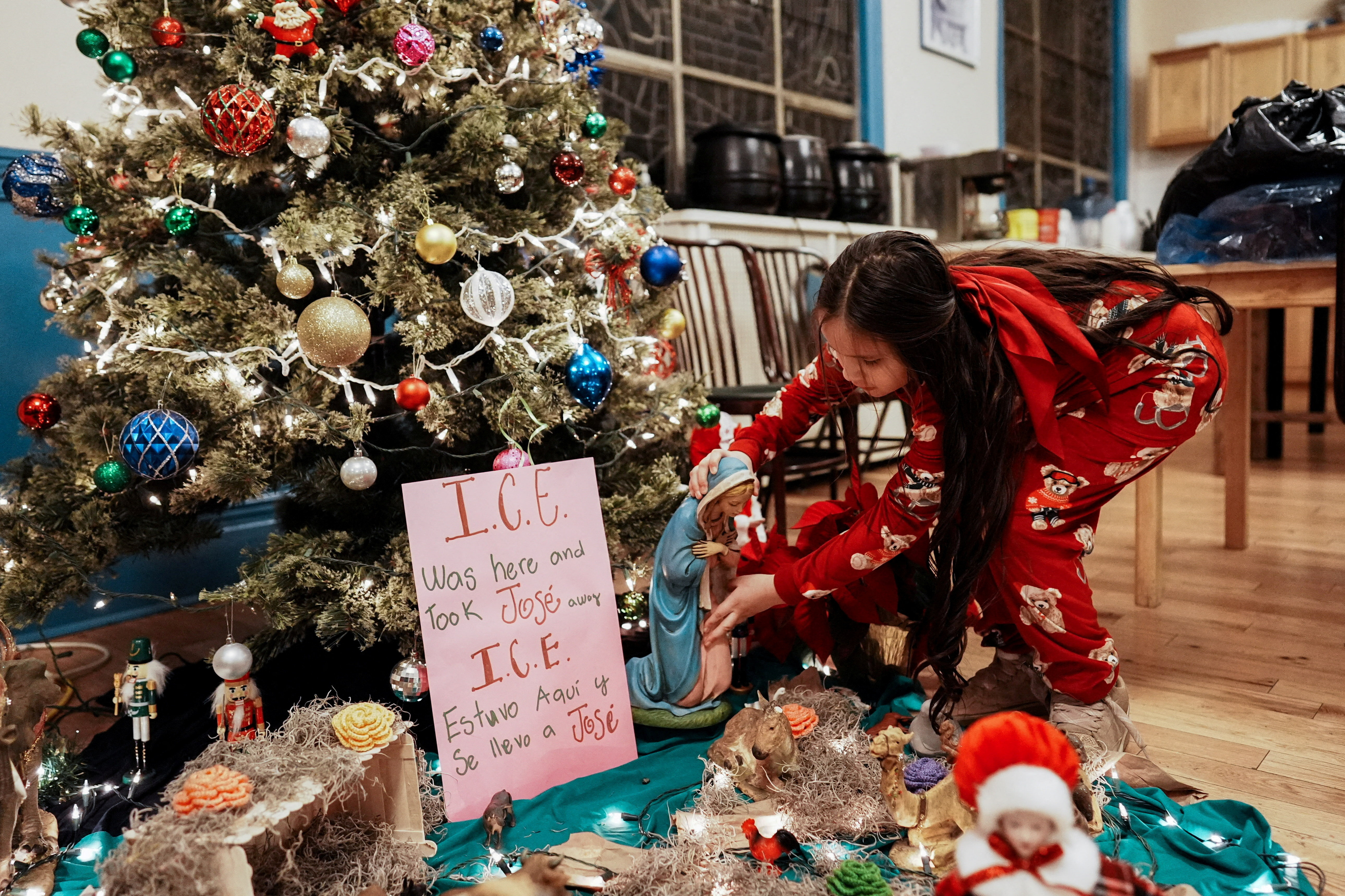 A little girl places an item below a Christmas tree that bears a sign explaining that ICE arrested a man named Jose there.