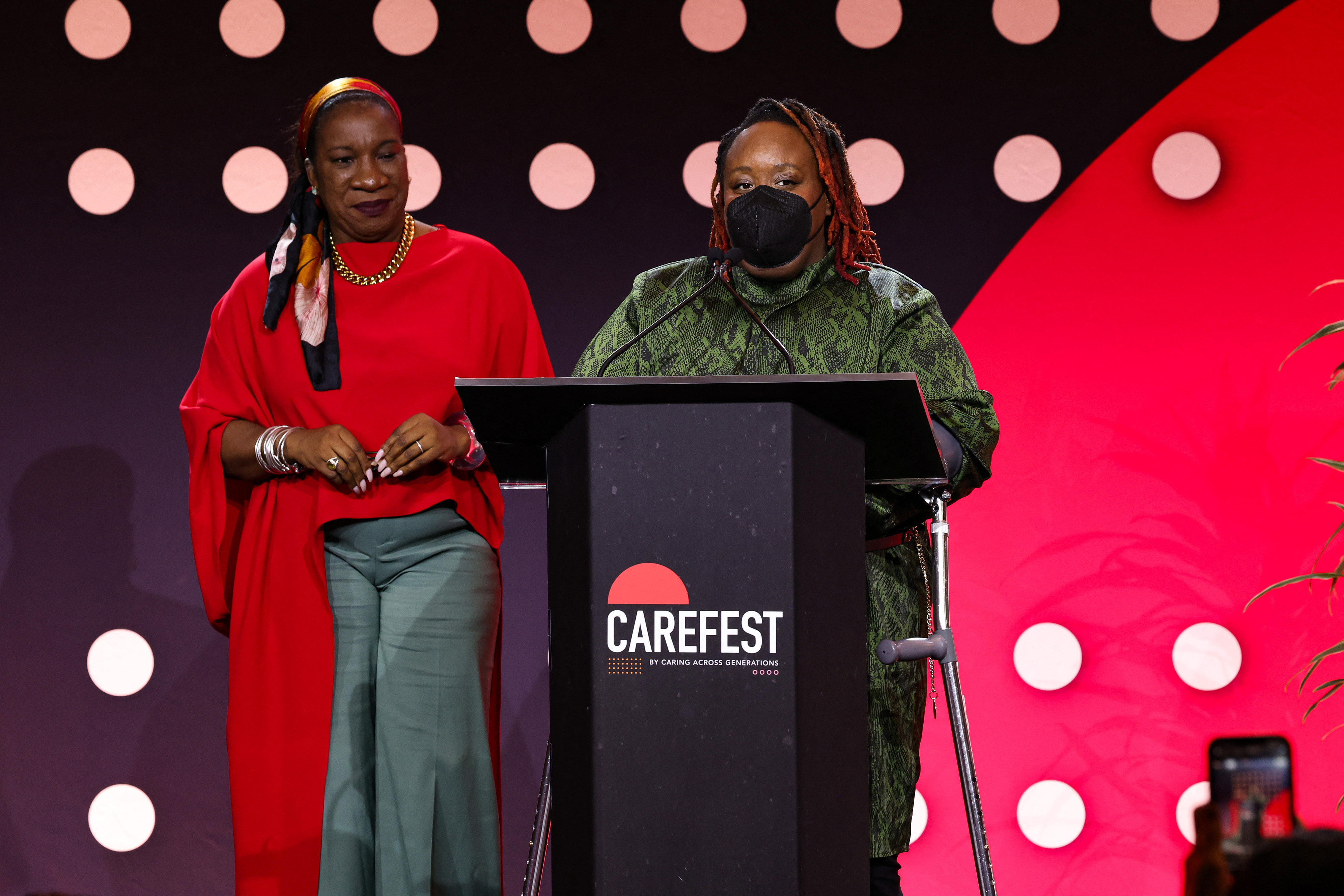 Tarana Burke and Imani Barbarin speak onstage during day one of CareFest. Imani stands at the podium in a green dress with a mask on.