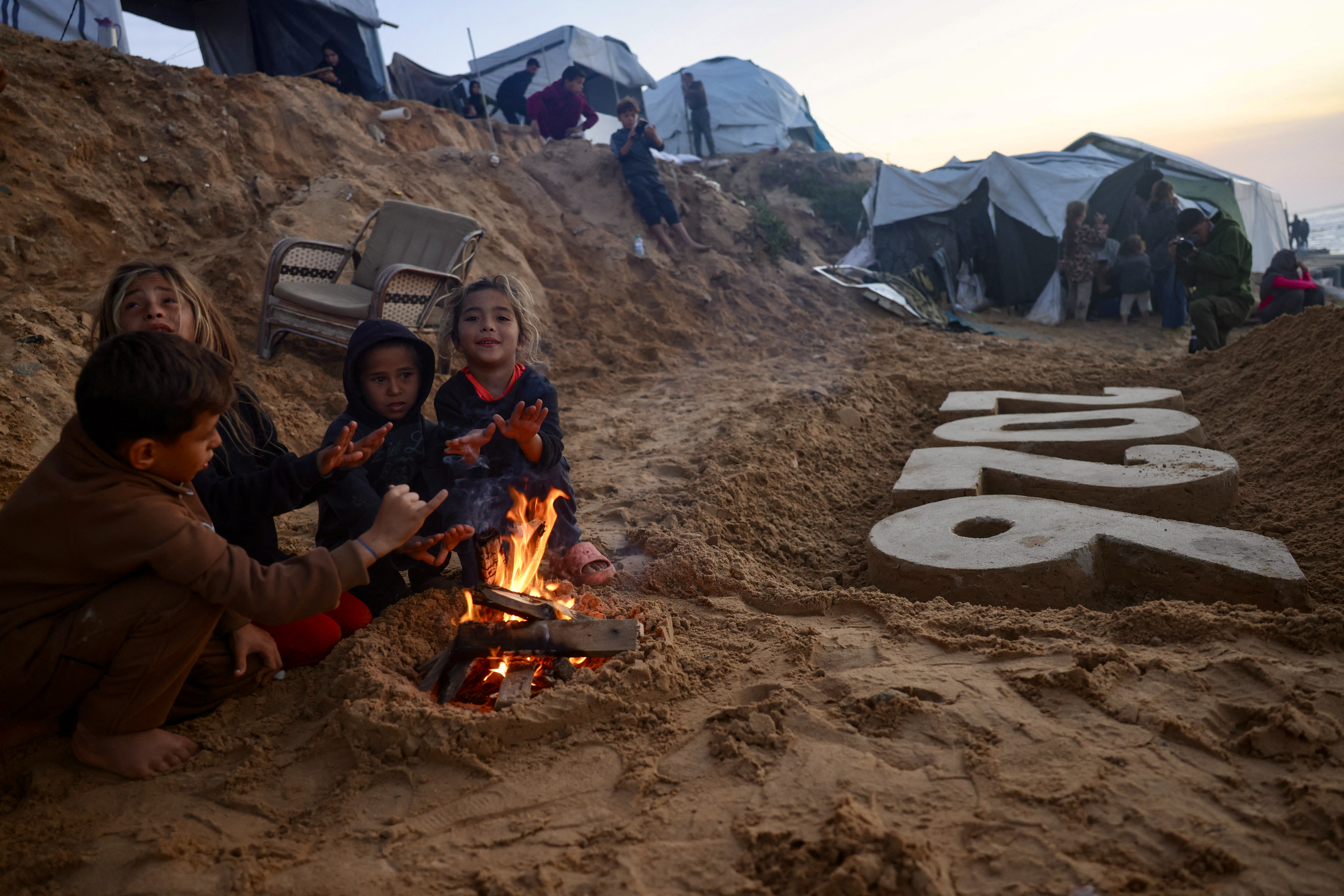 Children warm by the fire next to a sand sculpture by artist Yazed Abo Jarad of the coming year as Displaced Palestinians prepare to usher in the New Year in Deir El-Balah in the central Gaza Strip on December 30, 2025.
