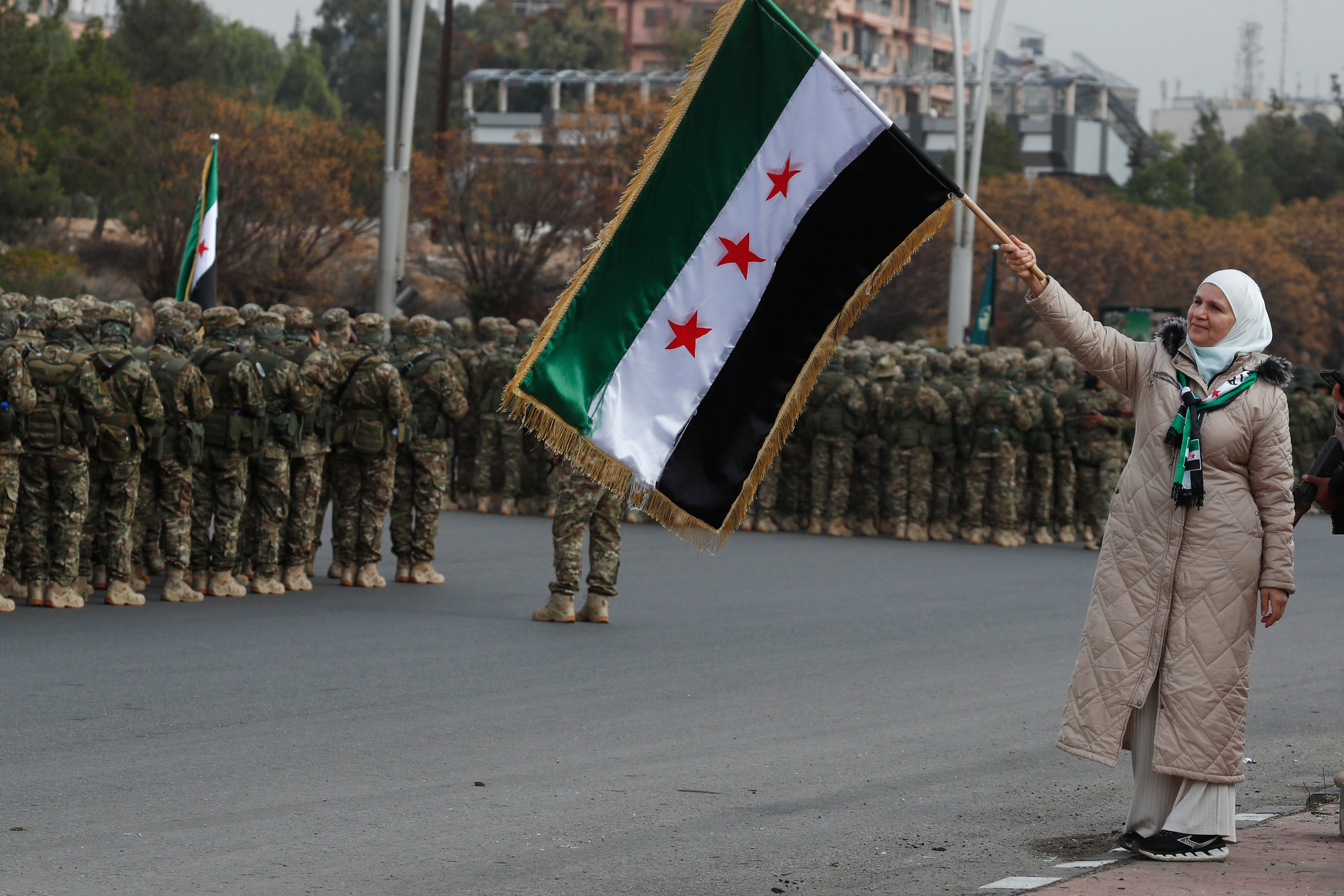 A woman waves the Syrian flag as members of the new Syrian army stand in formation during celebrations marking the first anniversary of the ousting of former President Bashar Assad in Damascus, Syria, Monday, Dec. 8, 2025. (AP Photo/Omar Sanadiki)