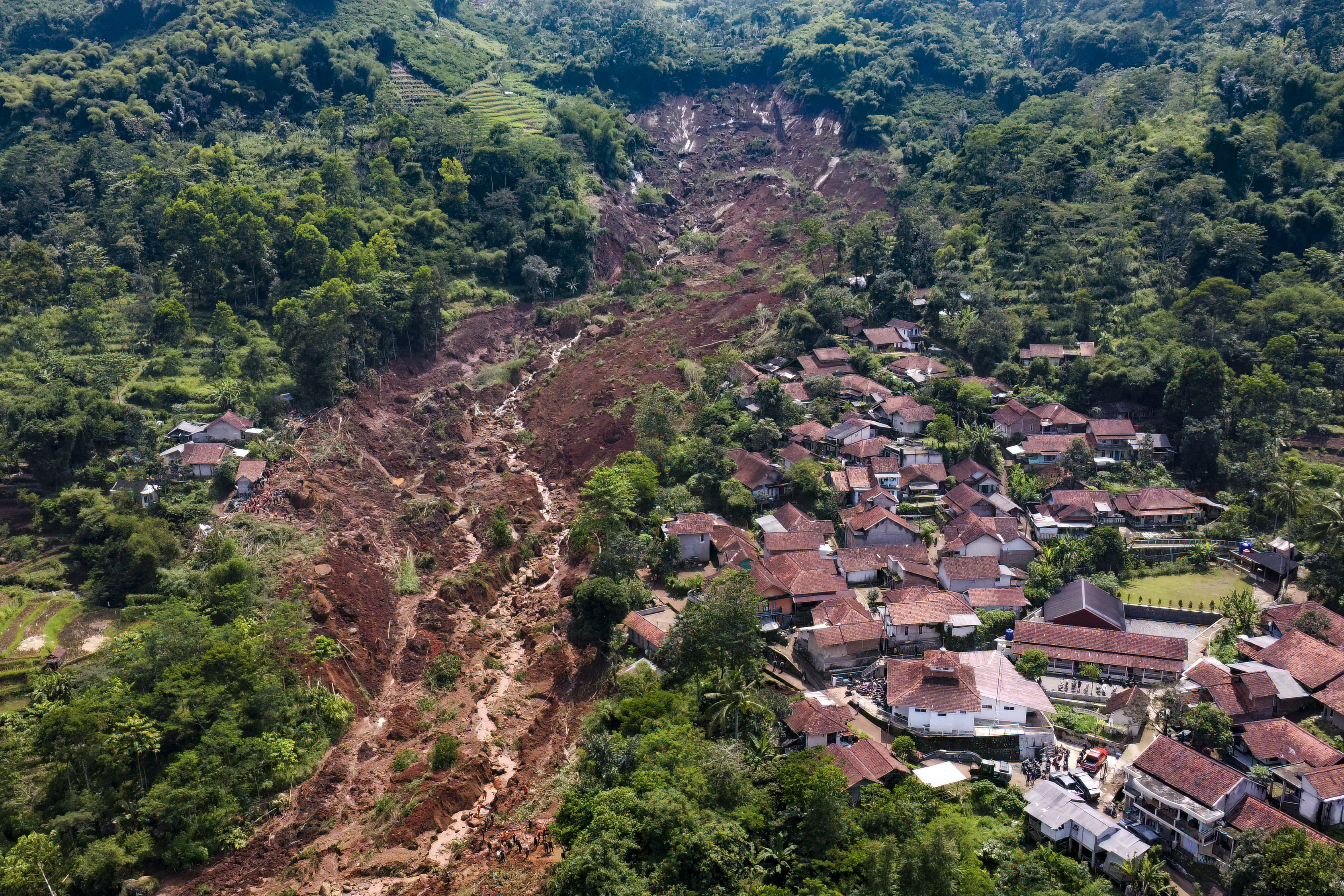 A view shows a brown landslide through a village surrounded by green.