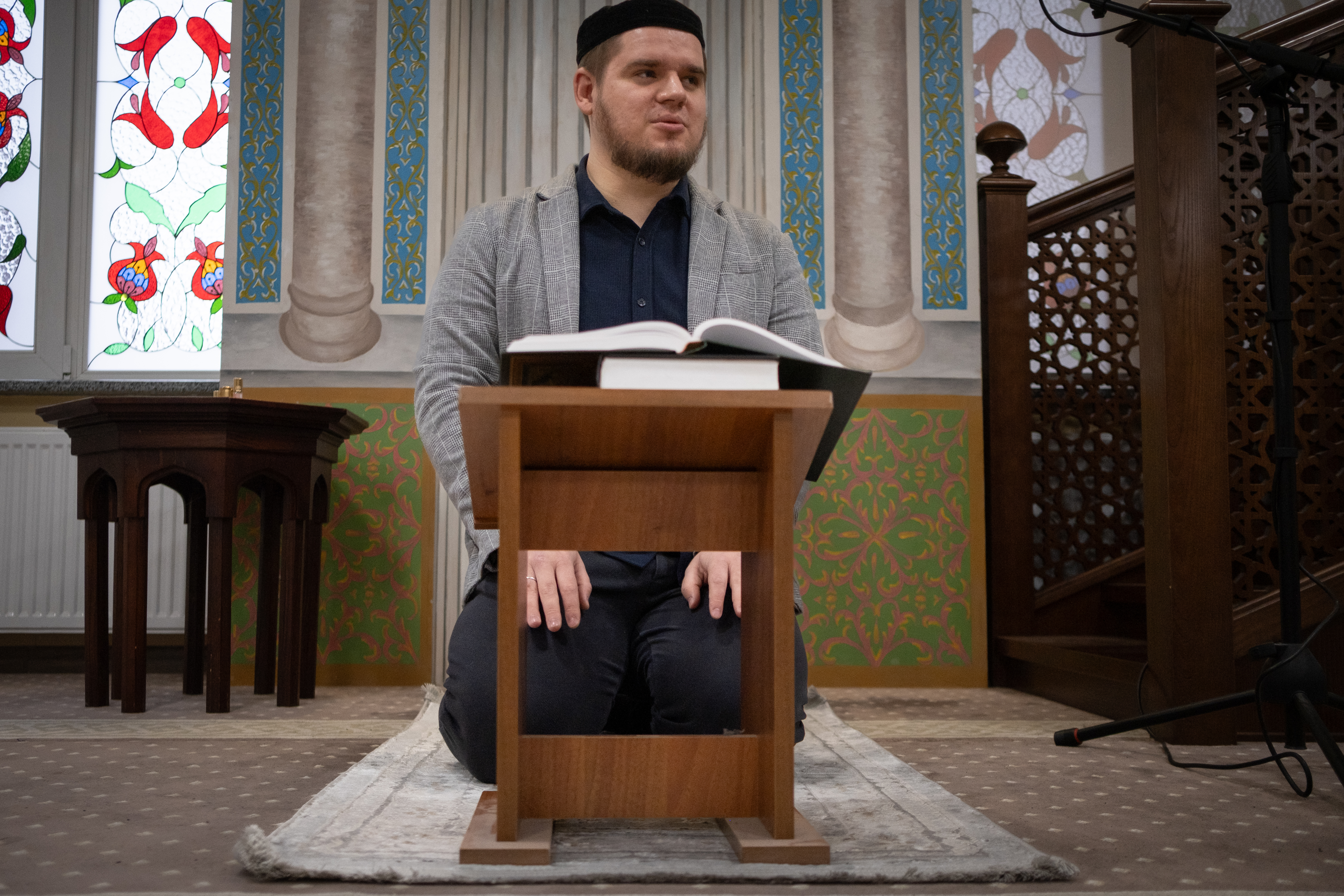 A man kneels on a carpet as he reads from a book.