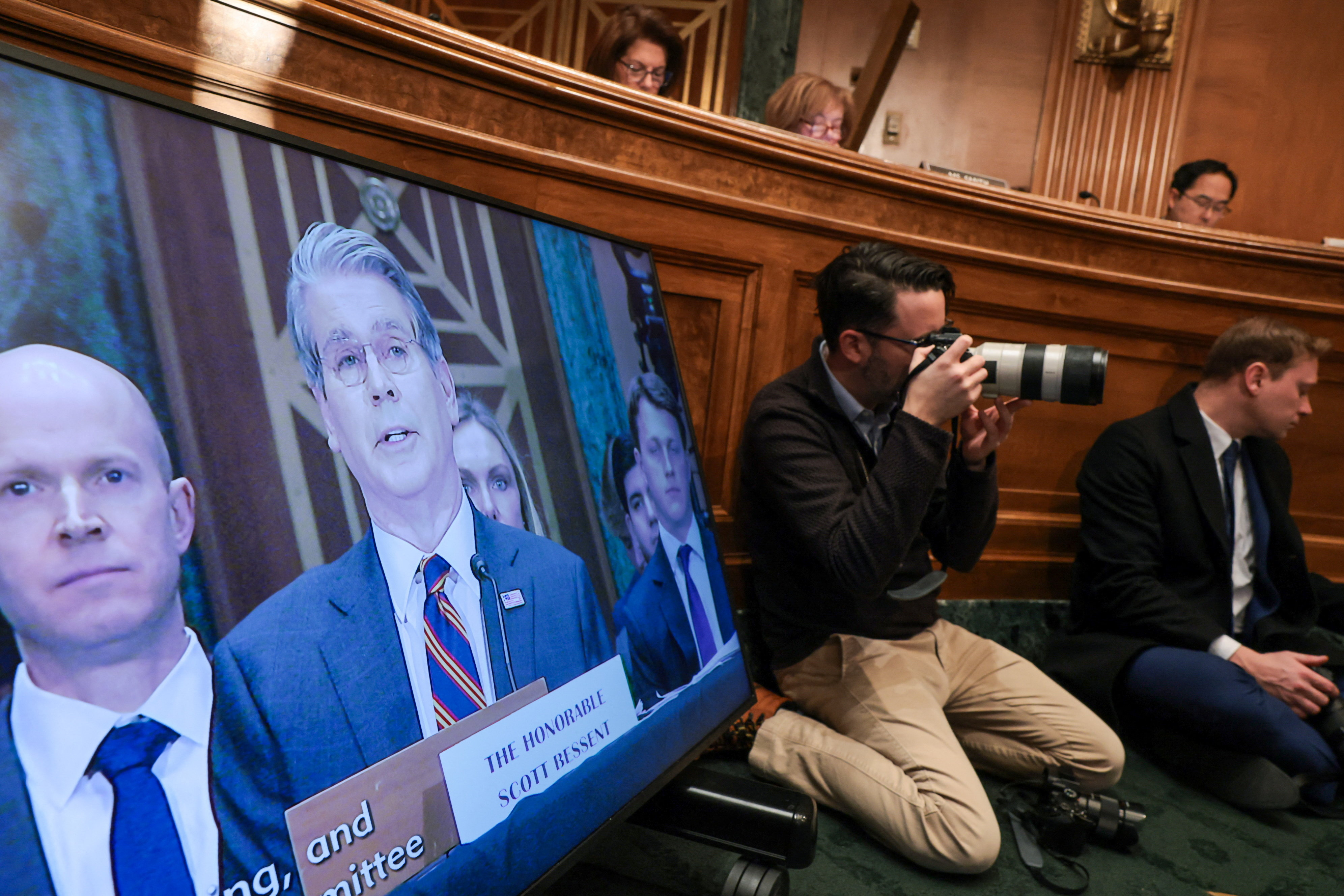 A screen shows Scott Bessent testifying at a Senate committee hearing. A photographer sits on the floor next to the screen.