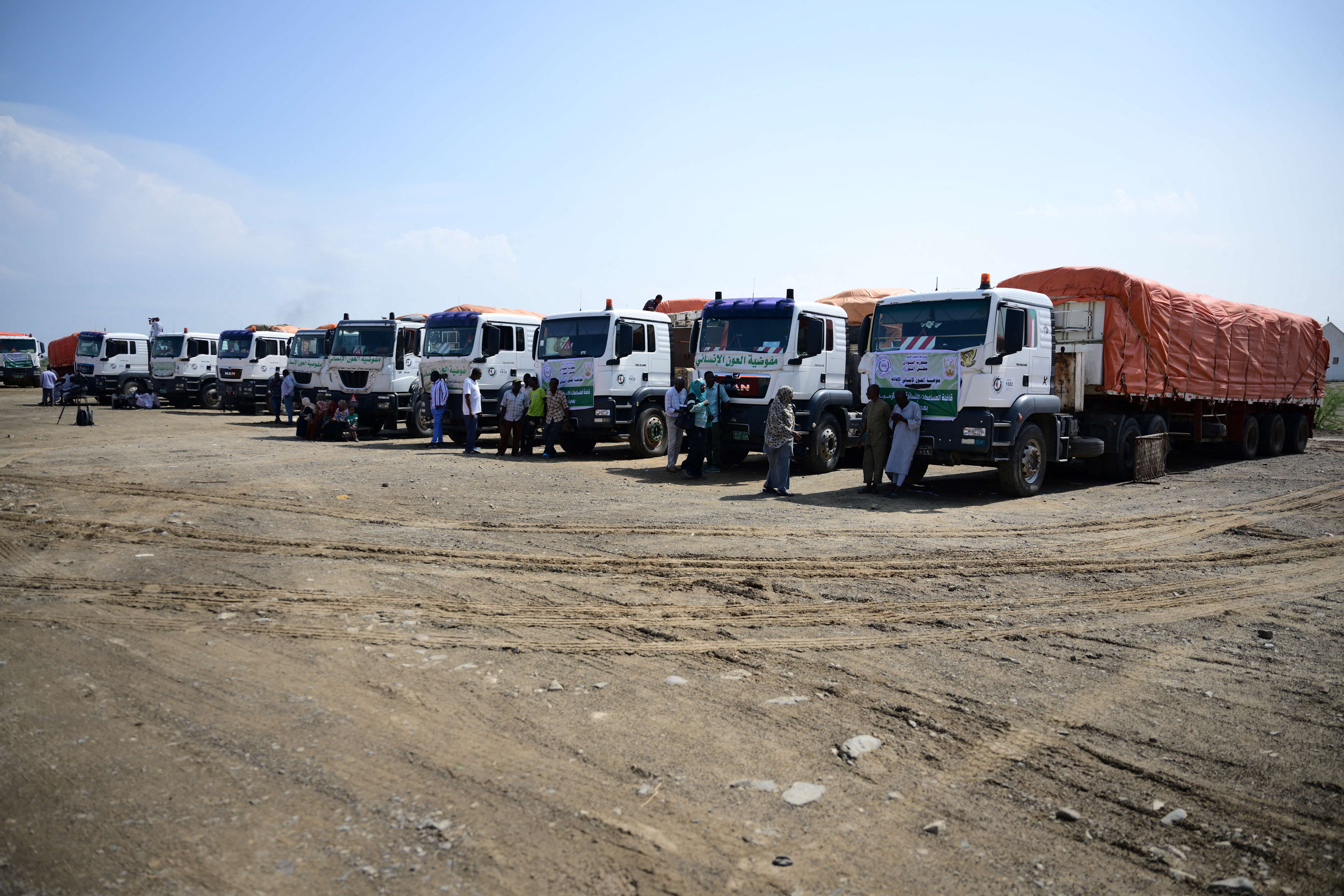 People stand near trucks loaded with aid in Port Sudan during the launch of a humanitarian aid convoy heading to the northern town of Al-Dabba, Sudan on November 13, 2025.