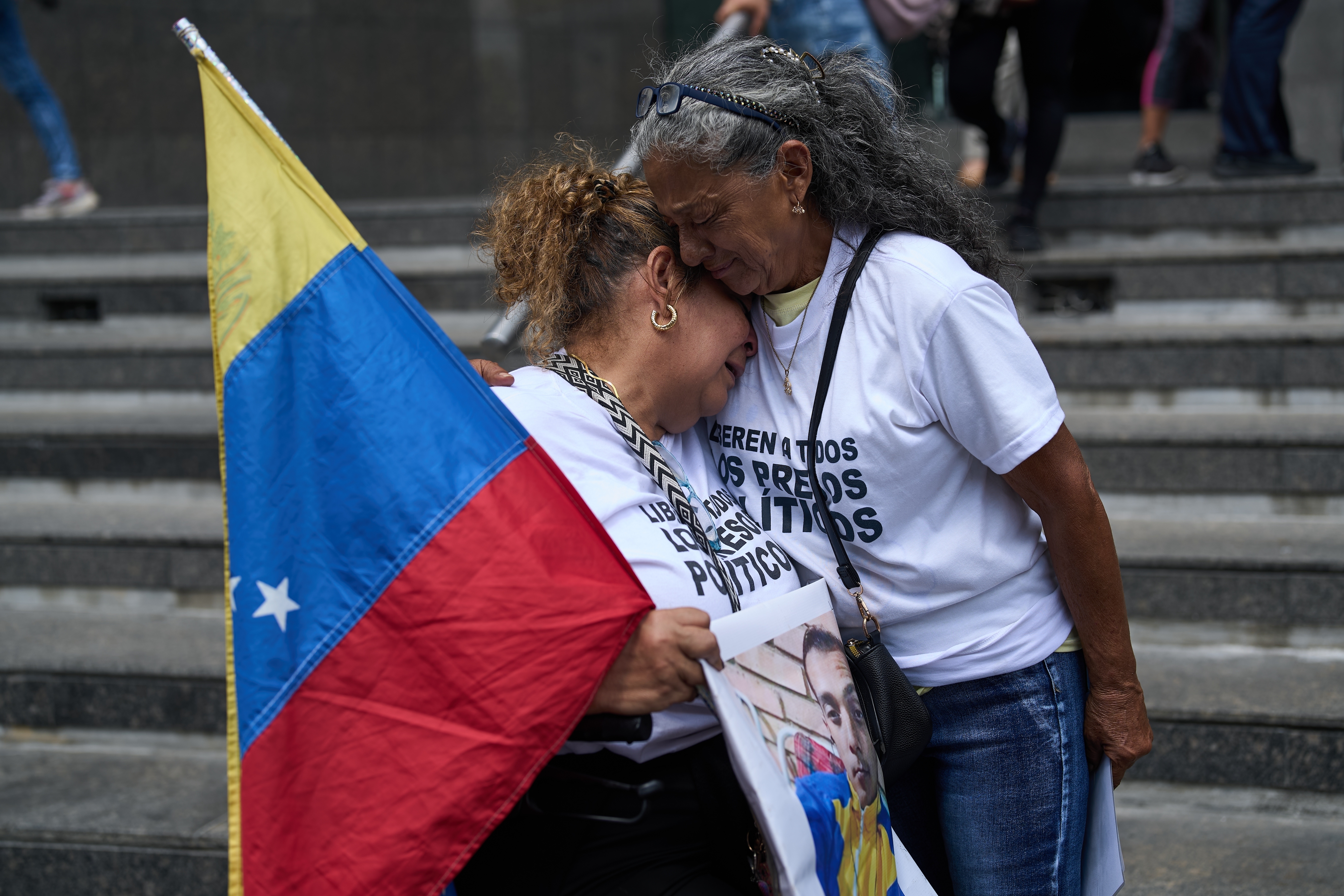 Barbara Bracho, left, mother of Gilberto Bracho, is embraced by Zoraida Gonzalez, mother of Miguel Estrada, both of whom consider their sons to be political prisoners, as they carry a Venezuelan flag