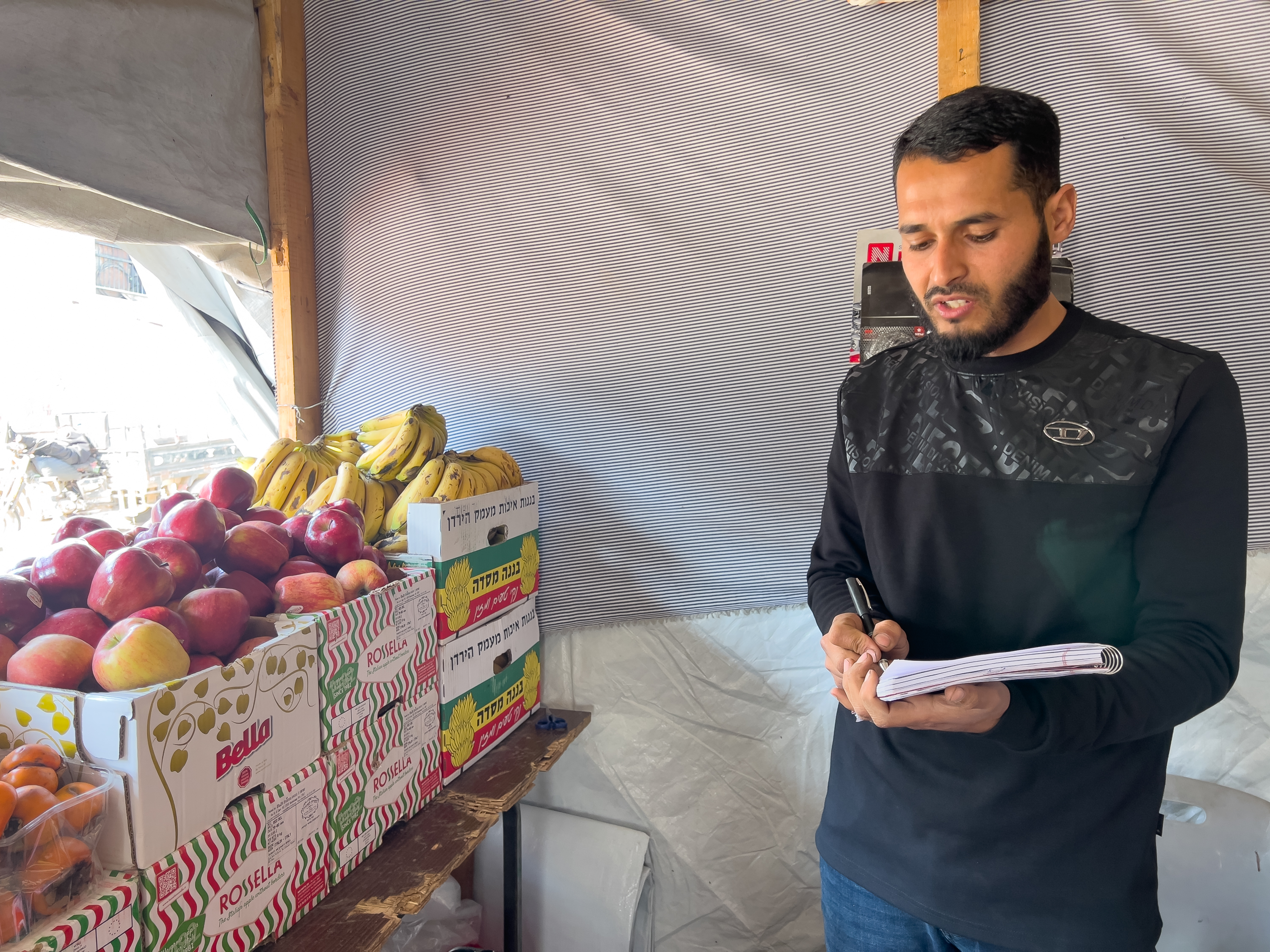 Abdallah Sukkar, owner of a street grocery stall, writing down customers' details in a notebook [Ola al-Asi/ Al Jazeera]