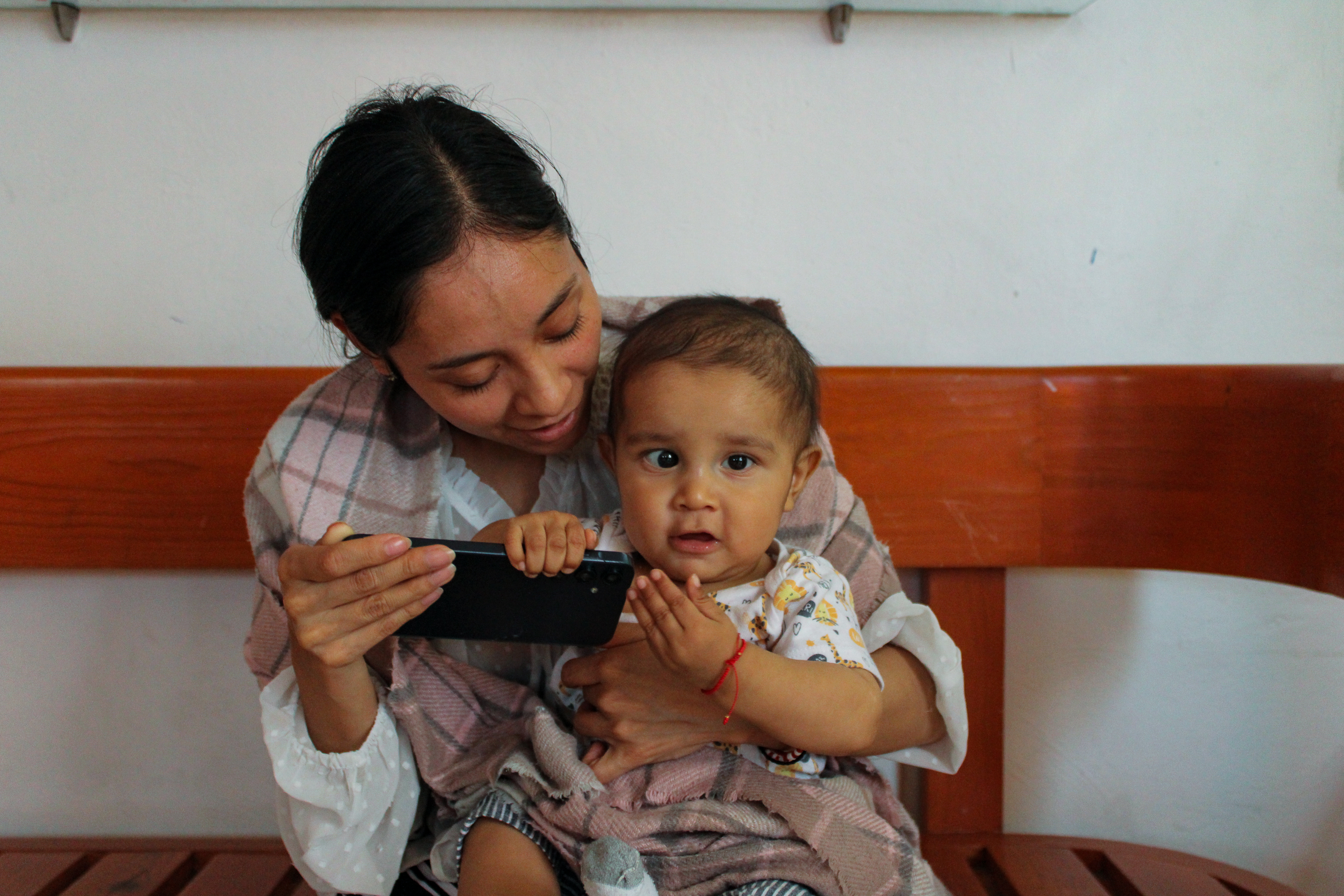 Luisa sits with Juan de Jesús on her knee in the lobby, playing his favourite nursery rhyme, La lechuza (“The Little Owl”), on her phone as they wait to be called_-1773831288