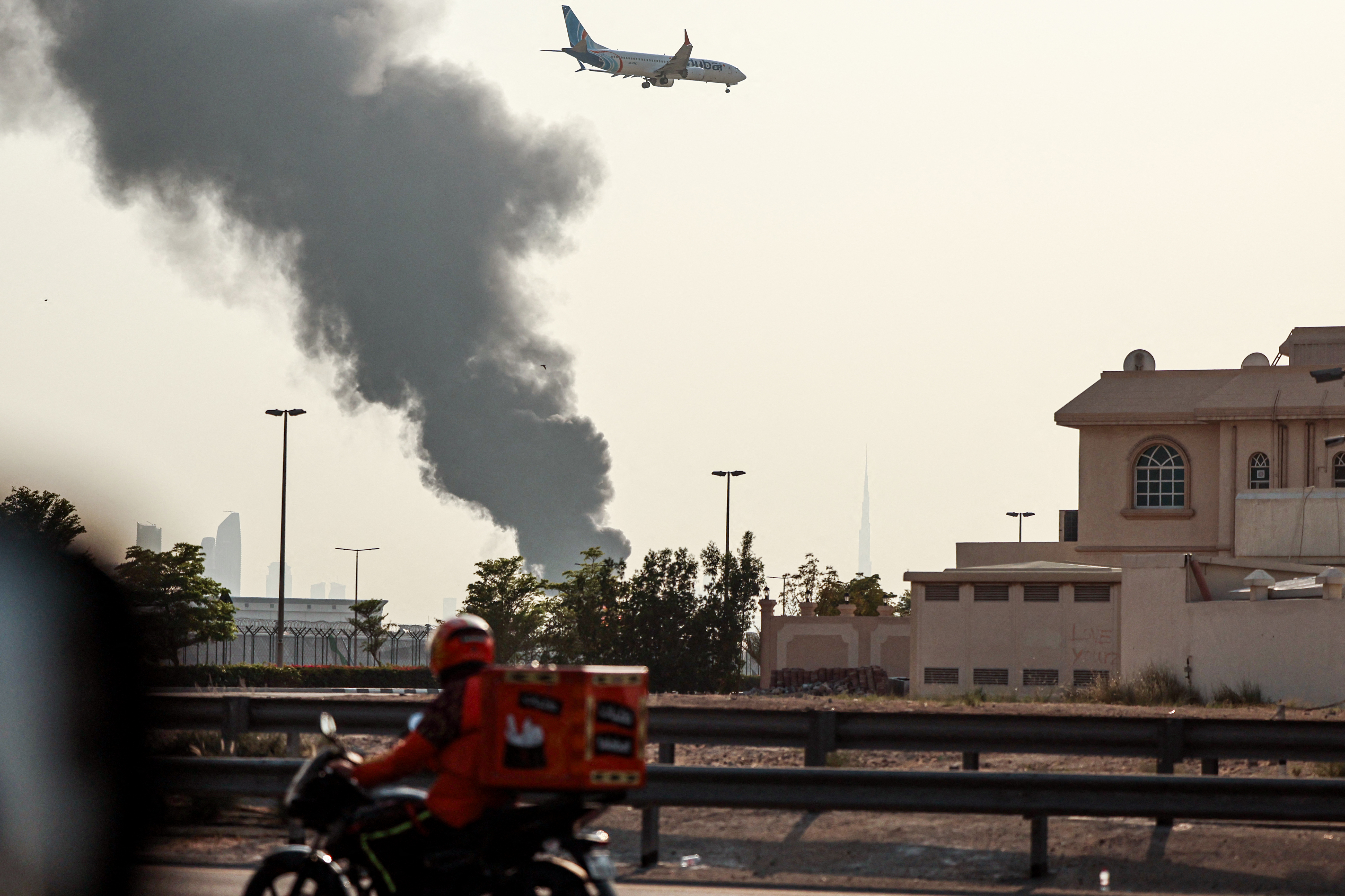 A delivery bike motors along a highway as an international low coast FlyDubai aircraft prepares for landing as a smoke plume rises from an ongoing fire near Dubai International Airport in Dubai on March 16, 2026.