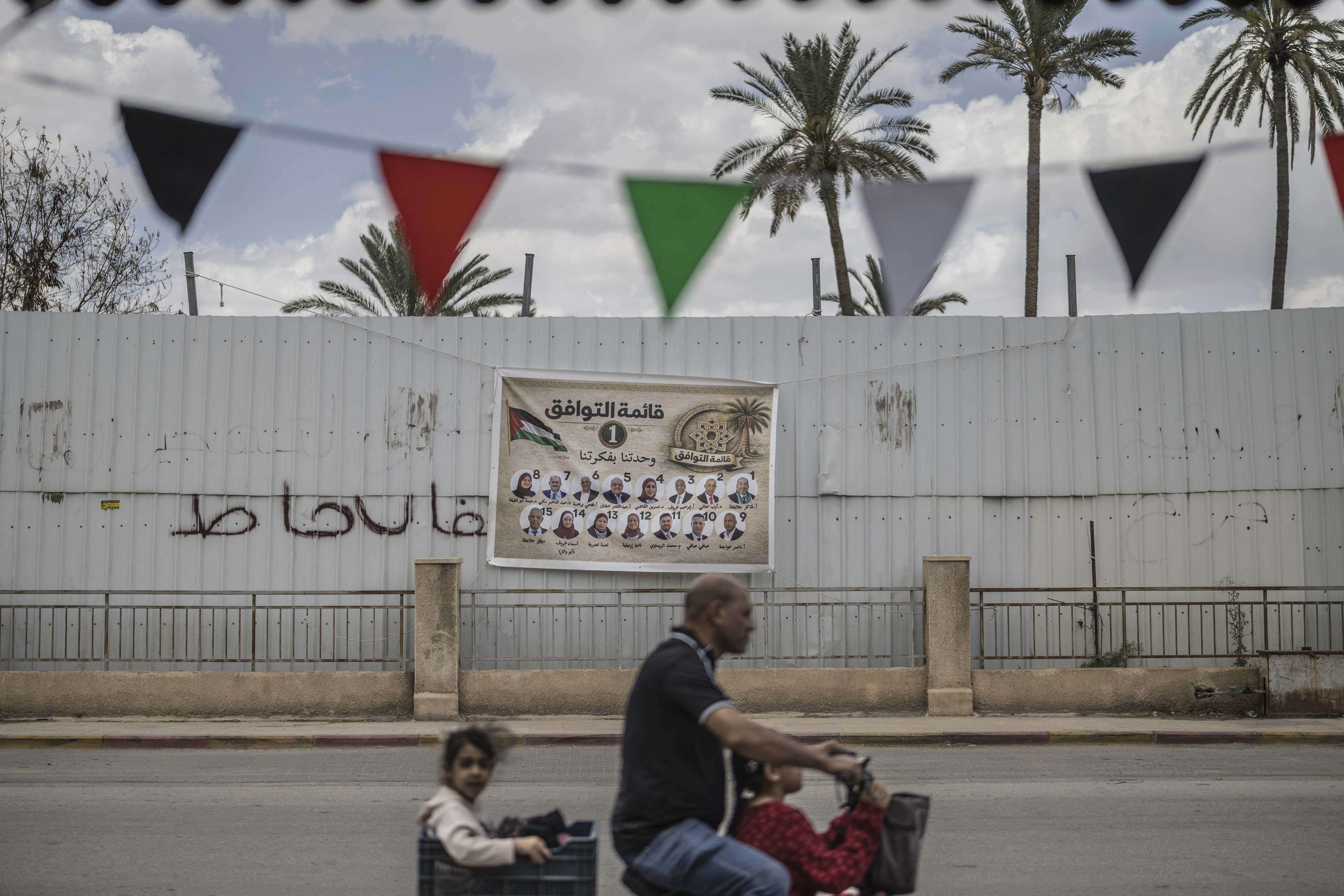 TOPSHOT - A man drives children on an electric bicycle past an election campaign banner showing candidates for the Jericho Governorate ahead of municipal elections, in the Israeli-occupied West Bank city of Jericho on April 20, 2026.
