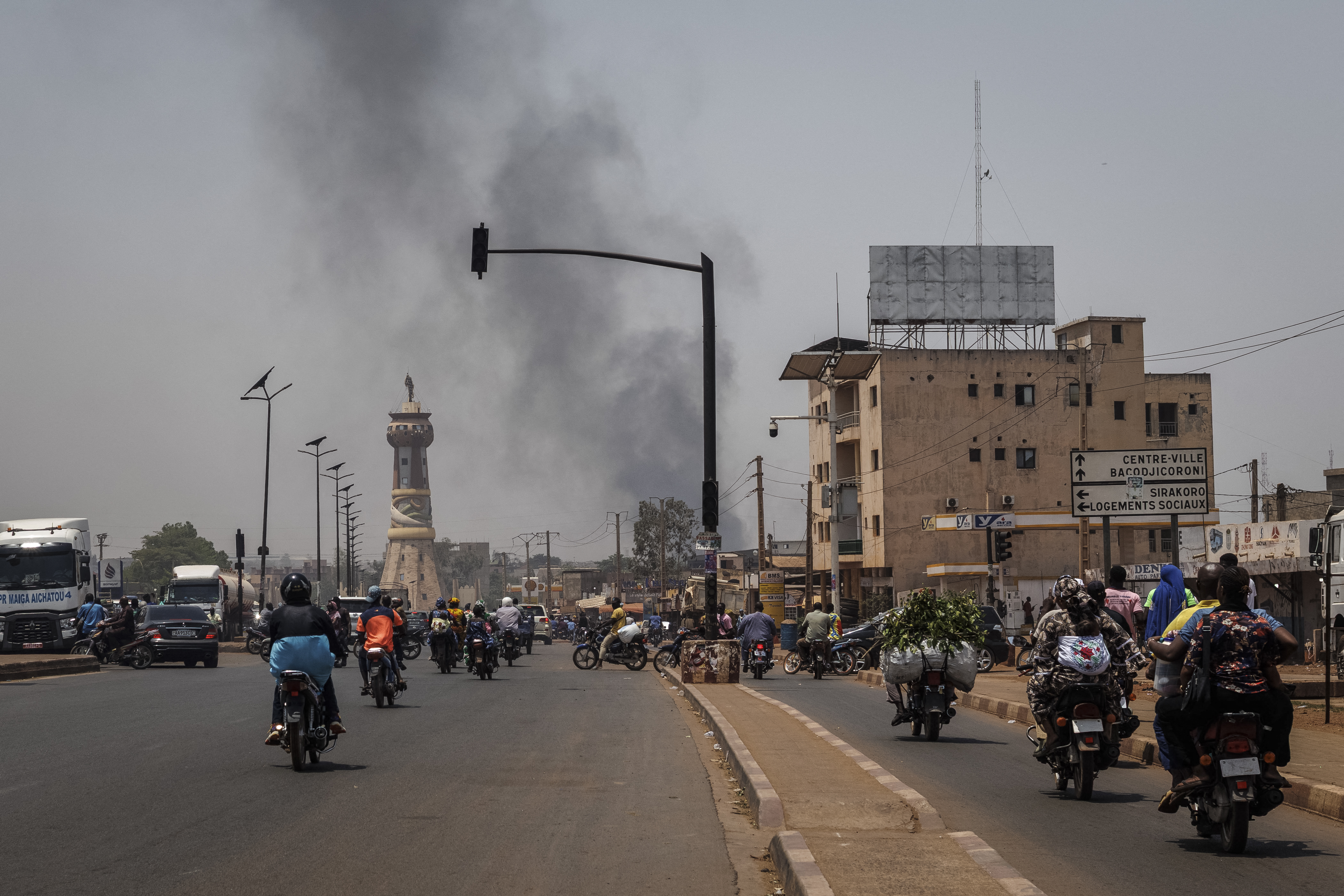 A column of black smoke rises above buildings as vehicles pass by the Africa Tower monument in Bamako, Mali