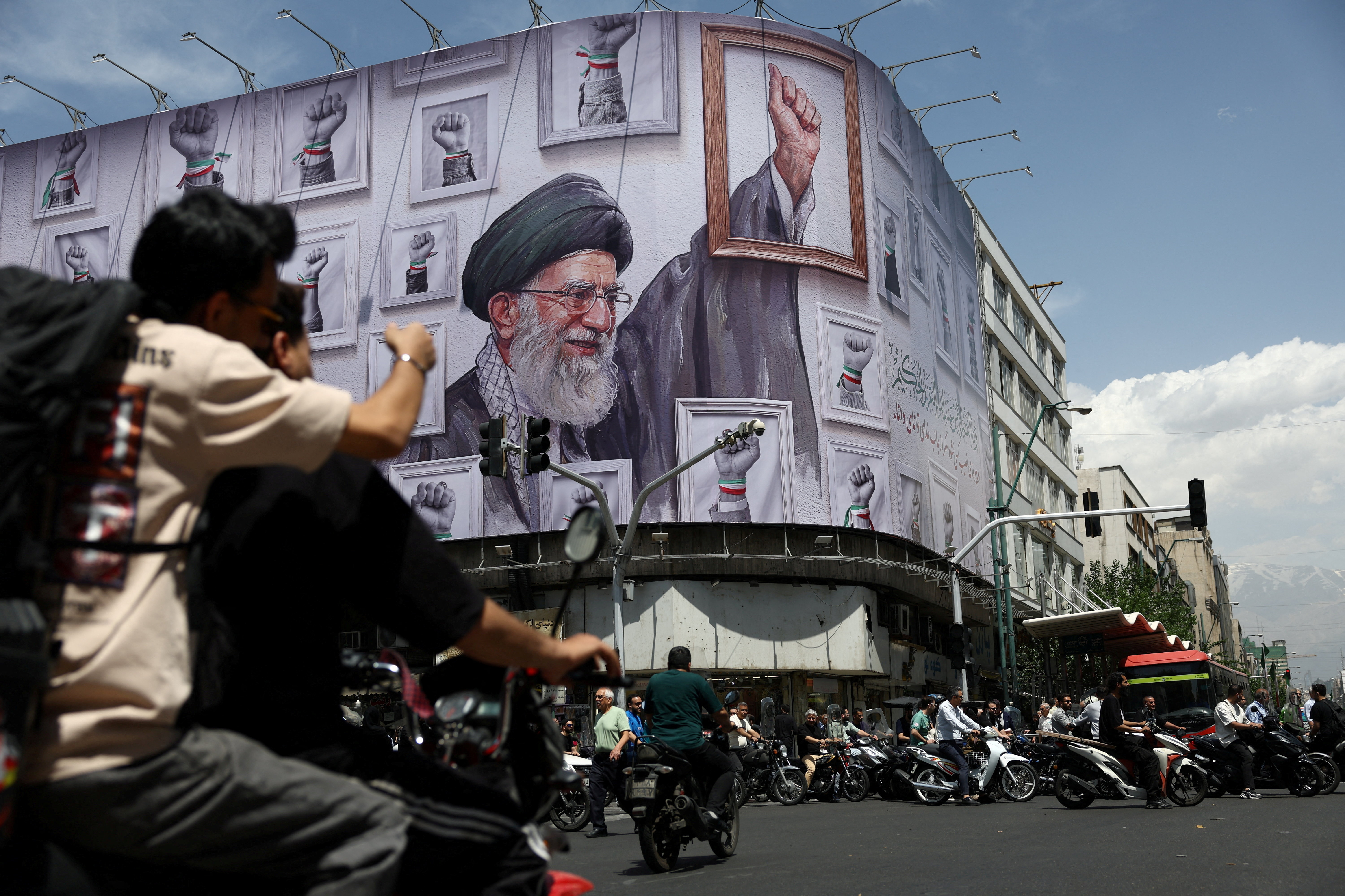 People ride motorcycles near a billboard featuring an image of the late supreme leader of Iran, Ayatollah Ali Khamenei.