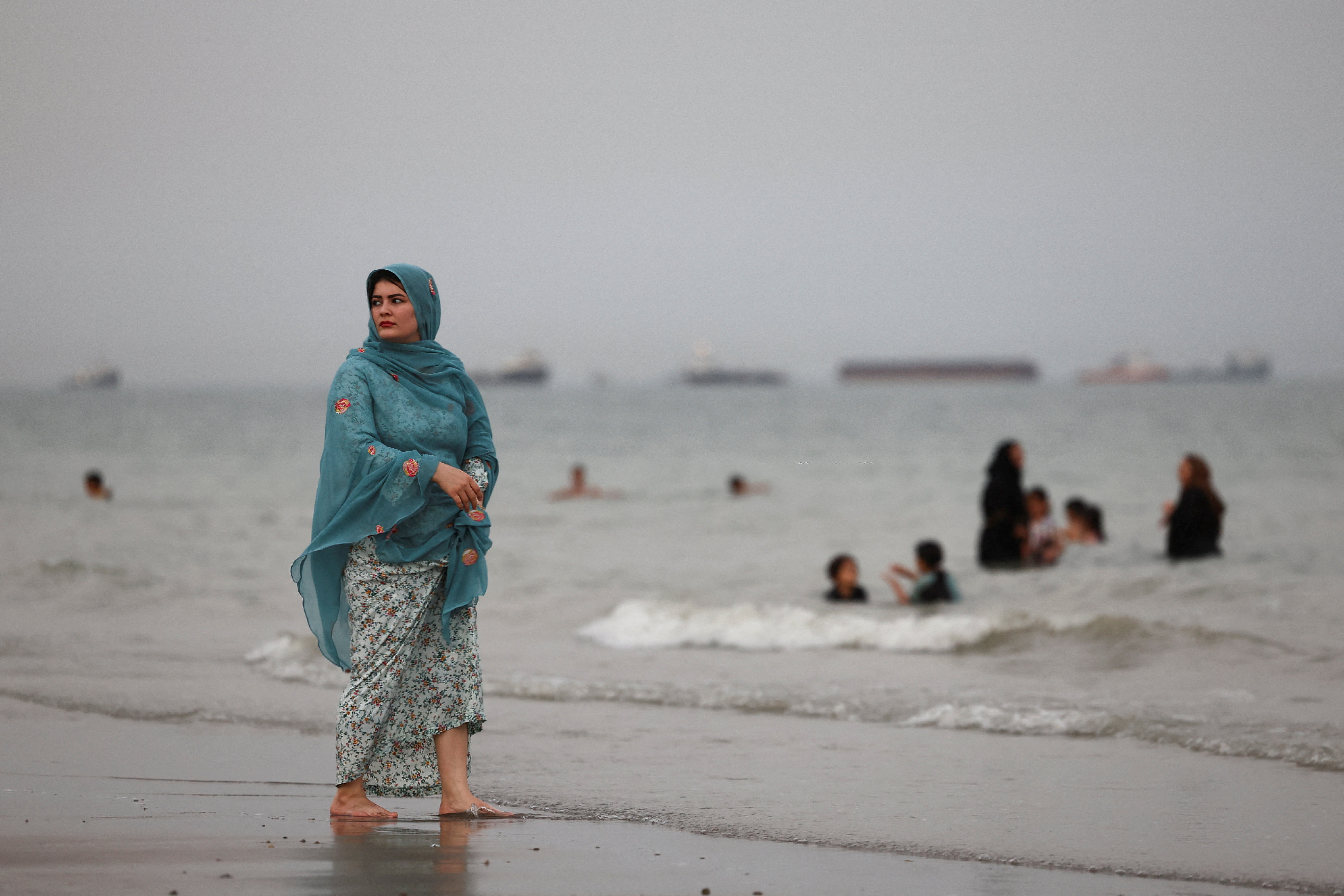 An Iranian woman walks on the beach as vessels in the Strait of Hormuz are visible near Bandar Abbas, Iran, on May 22, 2026