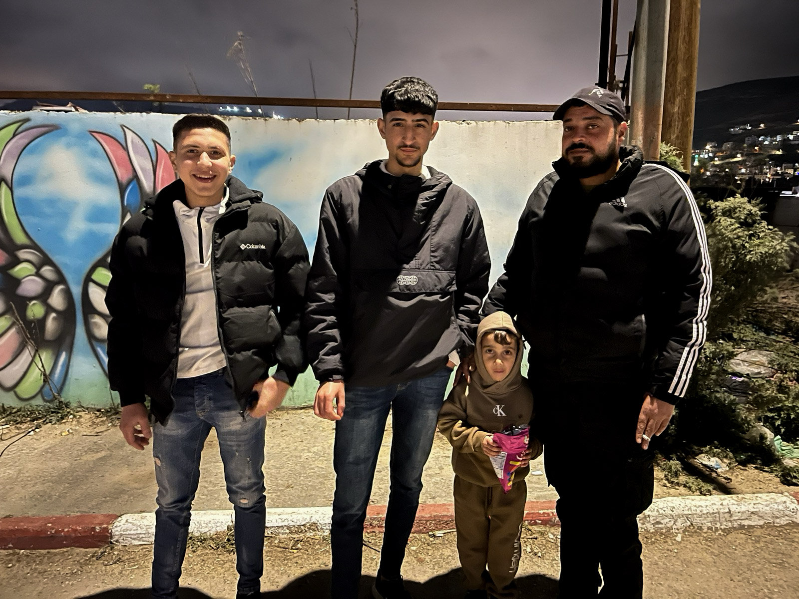 Yamen Habron, Amir Othman, and Islam Madani, along with Islam’s four-year-old son, at the entrance to Askar refugee camp in the occupied West Bank [Al Jazeera]