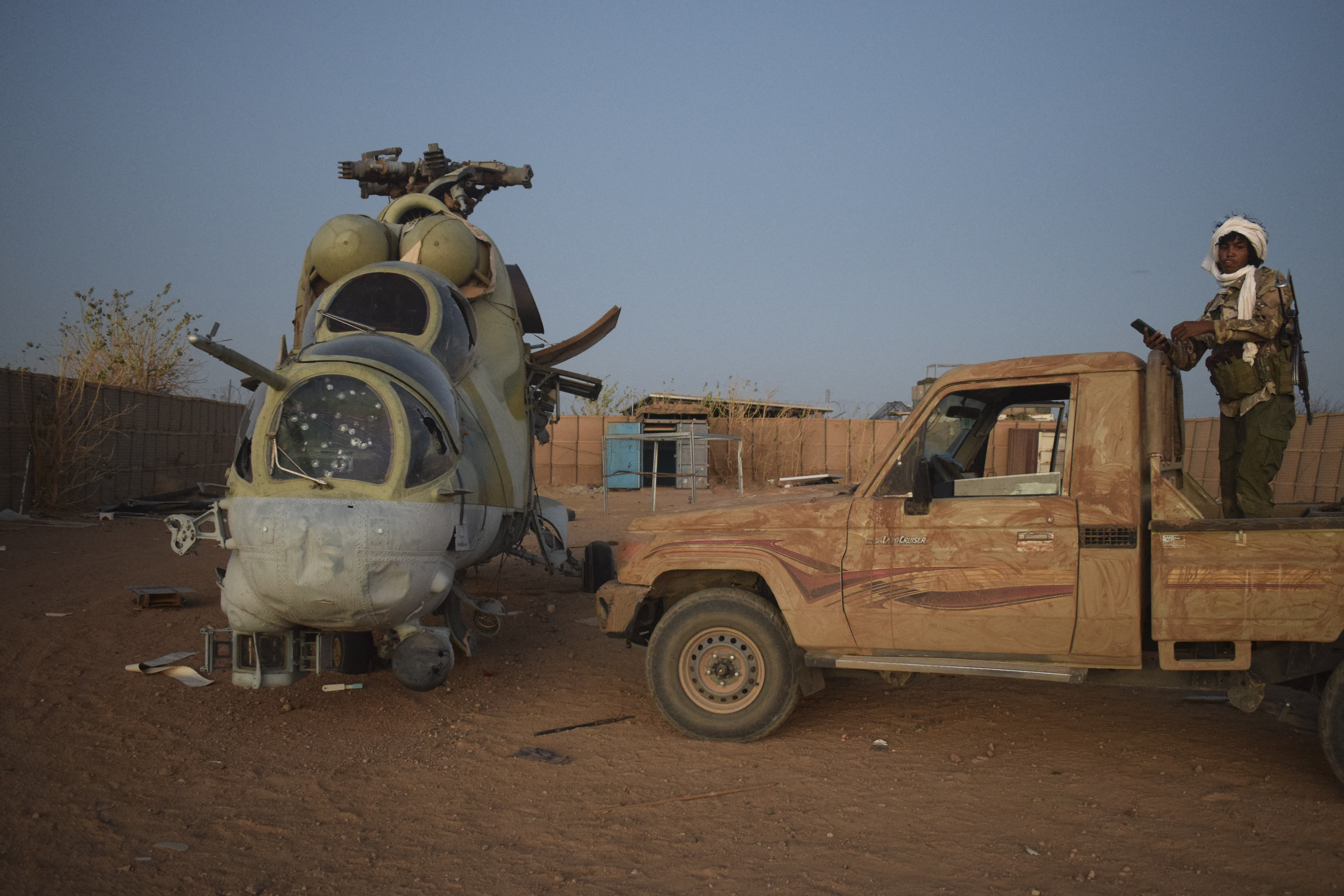 A soldier stands on a pick-up truck next to a damaged helicopter.