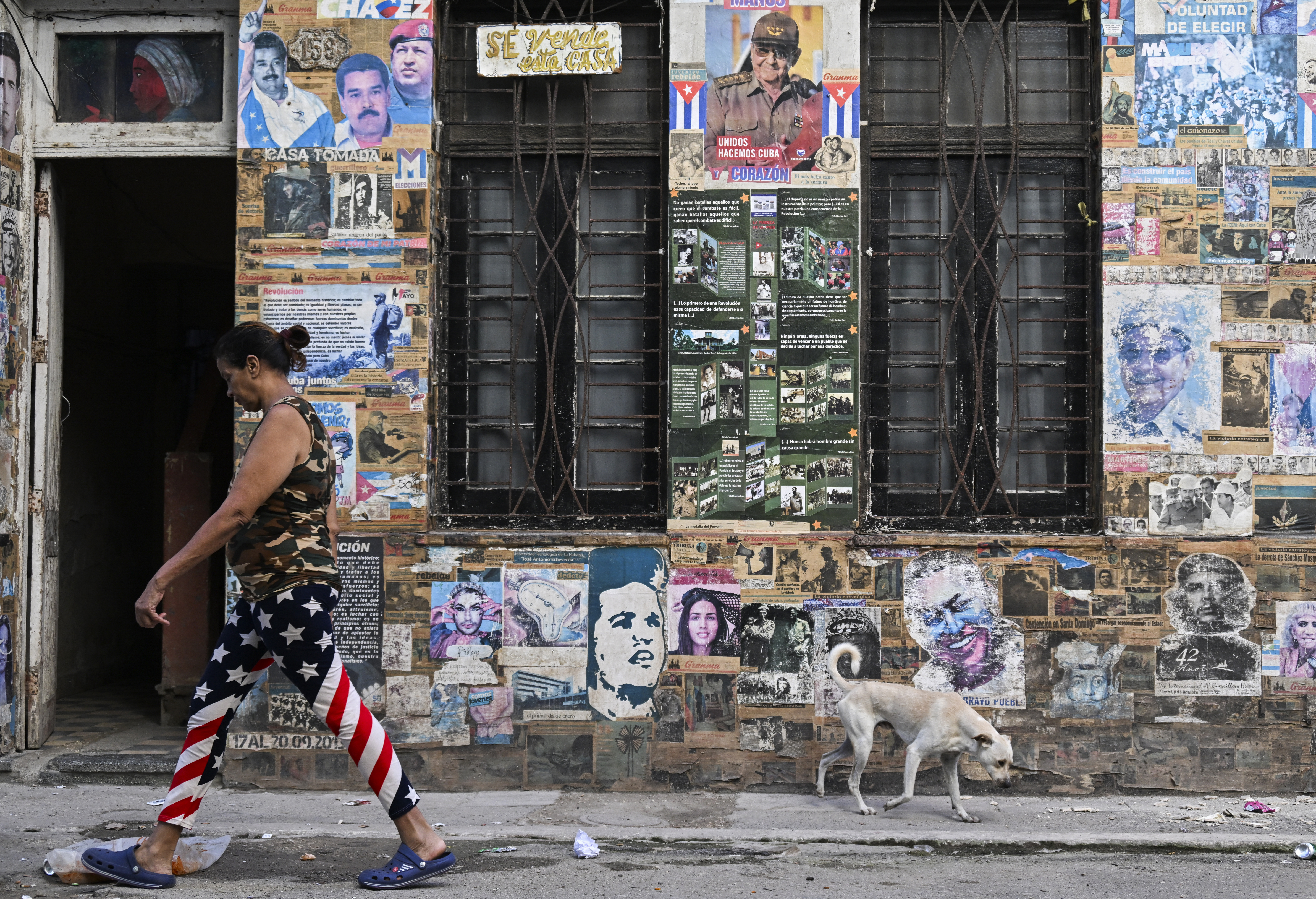 A woman wearing leggings bearing the flag of the United States, and a dog, walk past a house displaying a poster of the late Venezuela's leader Hugo Chavez, former Cuba's president Raul Castro and late revolutionary leader Argentine-born revolutionary leader Ernesto "Che" Guevara, in Havana, on May 19, 2026. (Photo by YAMIL LAGE / AFP)
