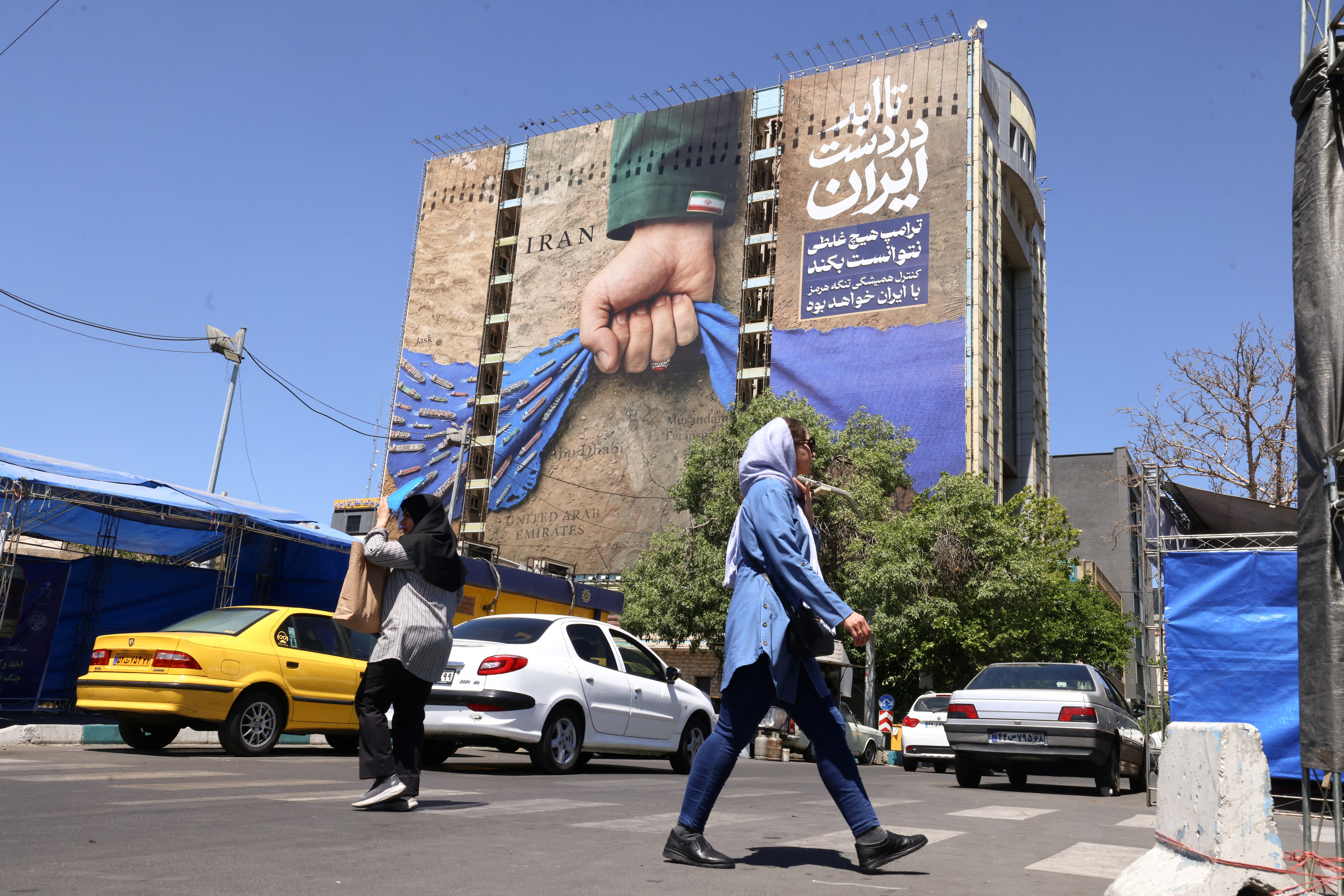 People cross a street near a billboard on the facade of a building depicting the Strait of Hormuz, with a caption in Persian reading 'Forever in Iran’s Hand'.