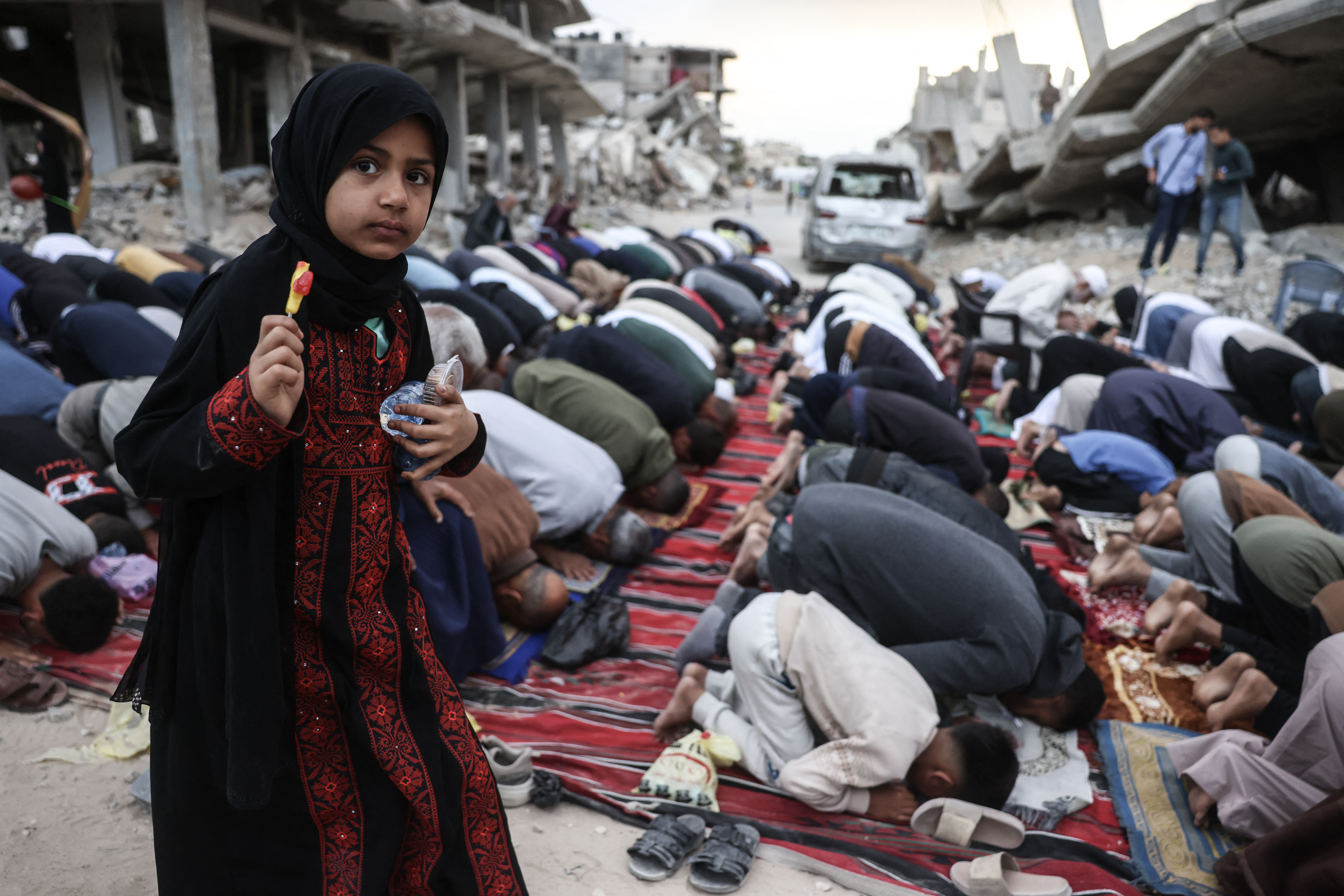 A girl holding a piece of candy stands among Palestinians performing morning prayers marking the start of Eid al-Adha, the Feast of Sacrifice, on a heavily damaged street in Khan Yunis in the southern Gaza Strip on May 27, 2026.