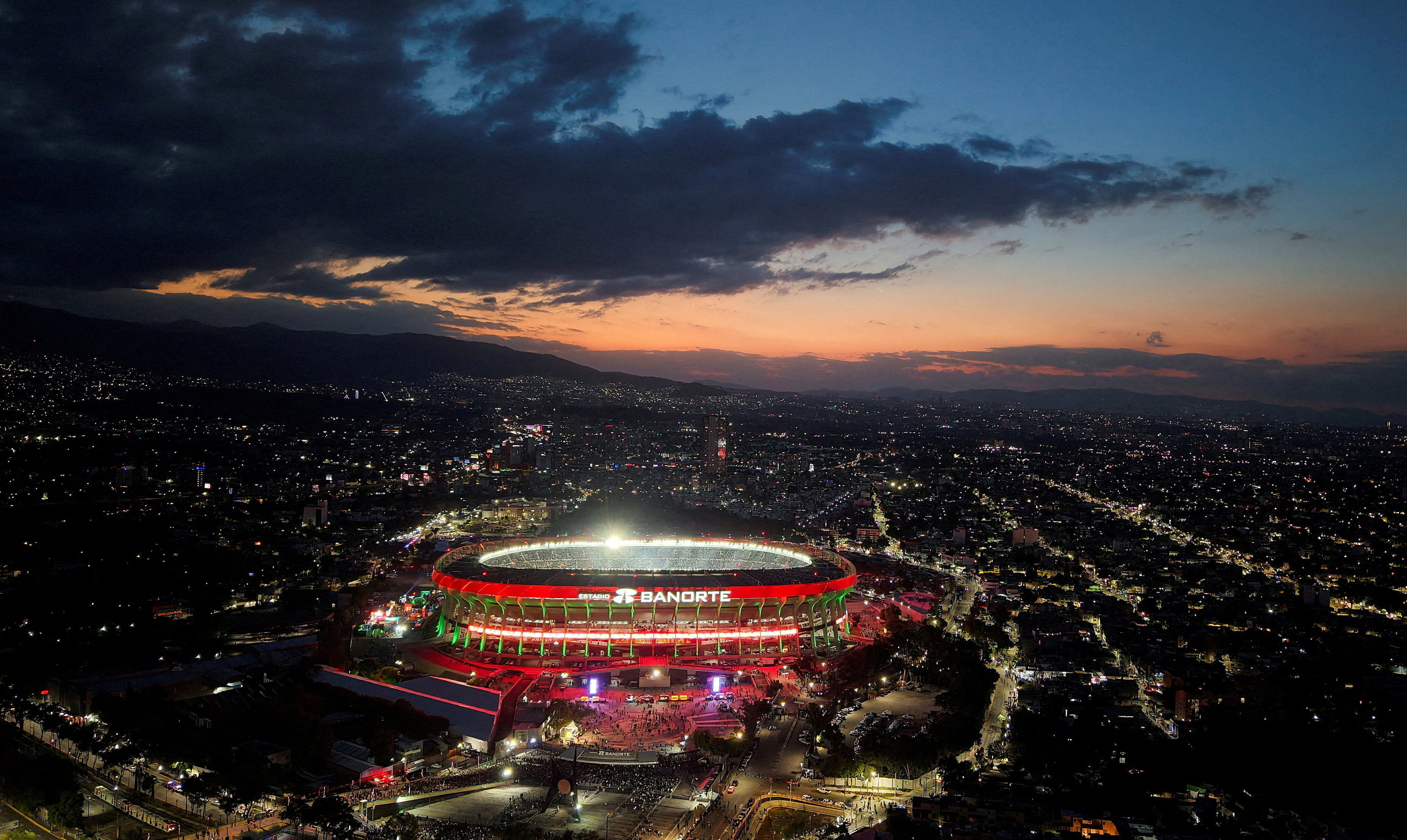 Azteca Stadium lit up as the sun sets with dark clouds overhead