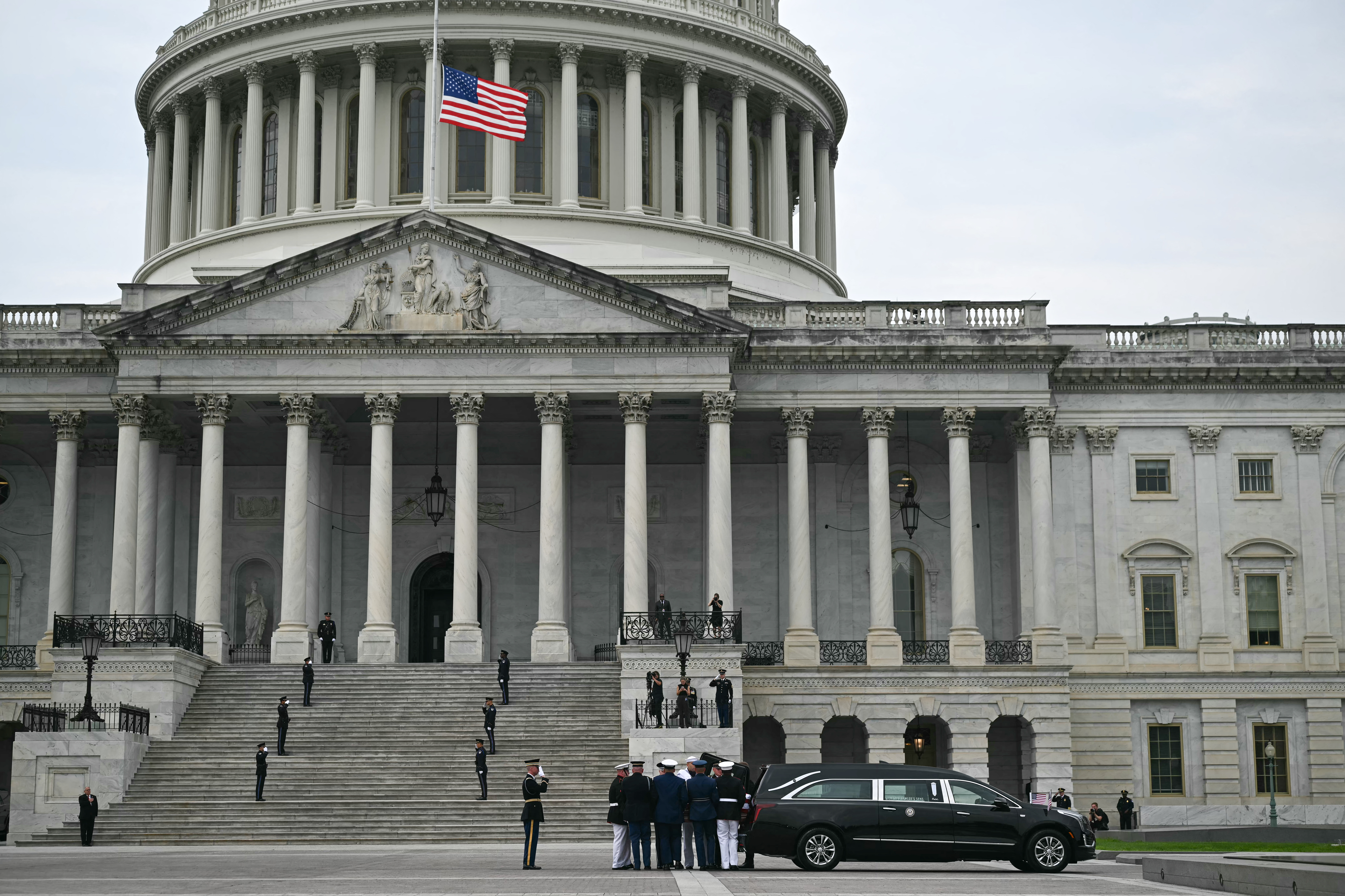 Uniformed service members carry a flag-draped casket down a set of steps.