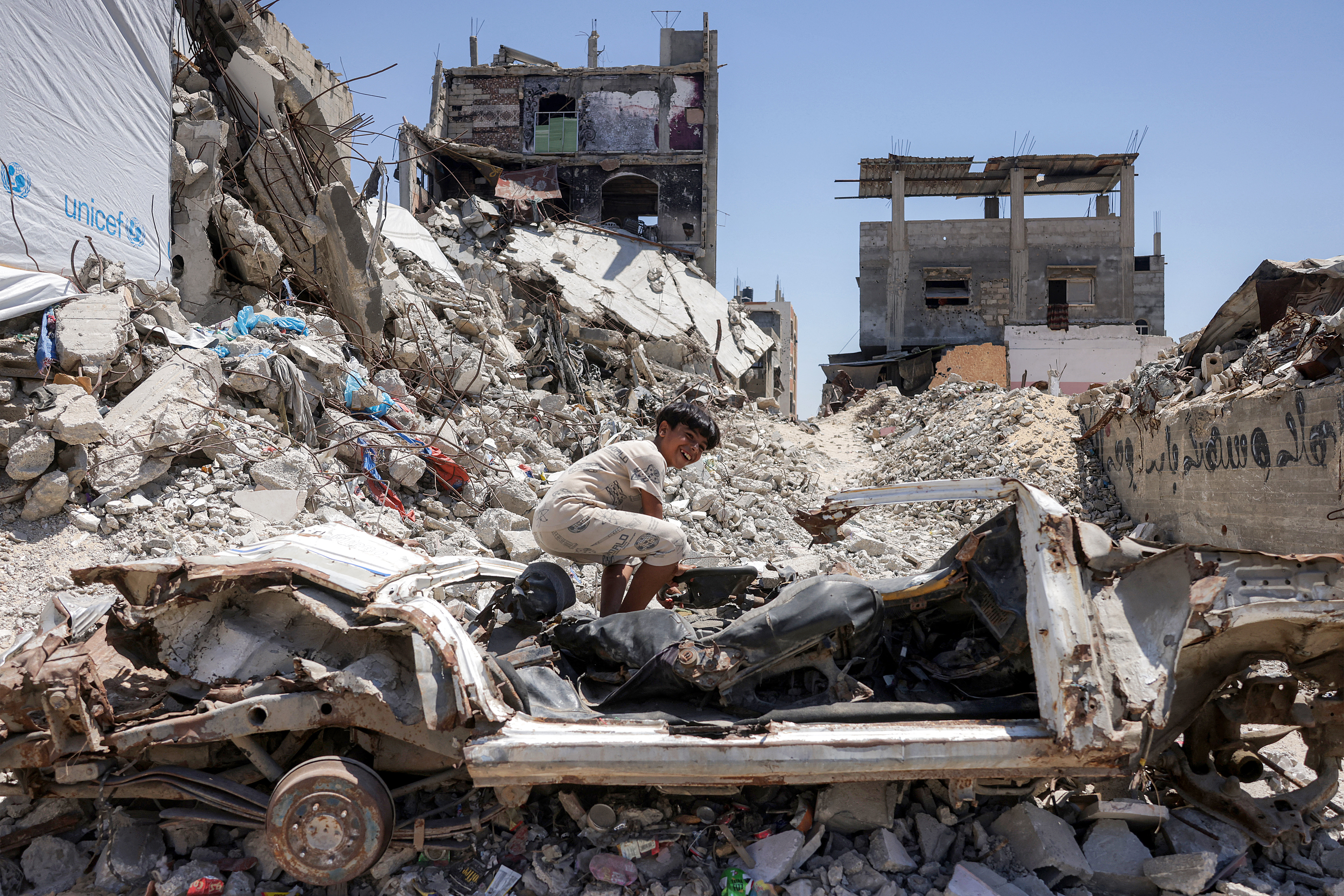 A boy sits on the frame of a destroyed vehicle near the rubble of collapsed buildings in Khan Yunis in the southern Gaza Strip on August 25, 2026.