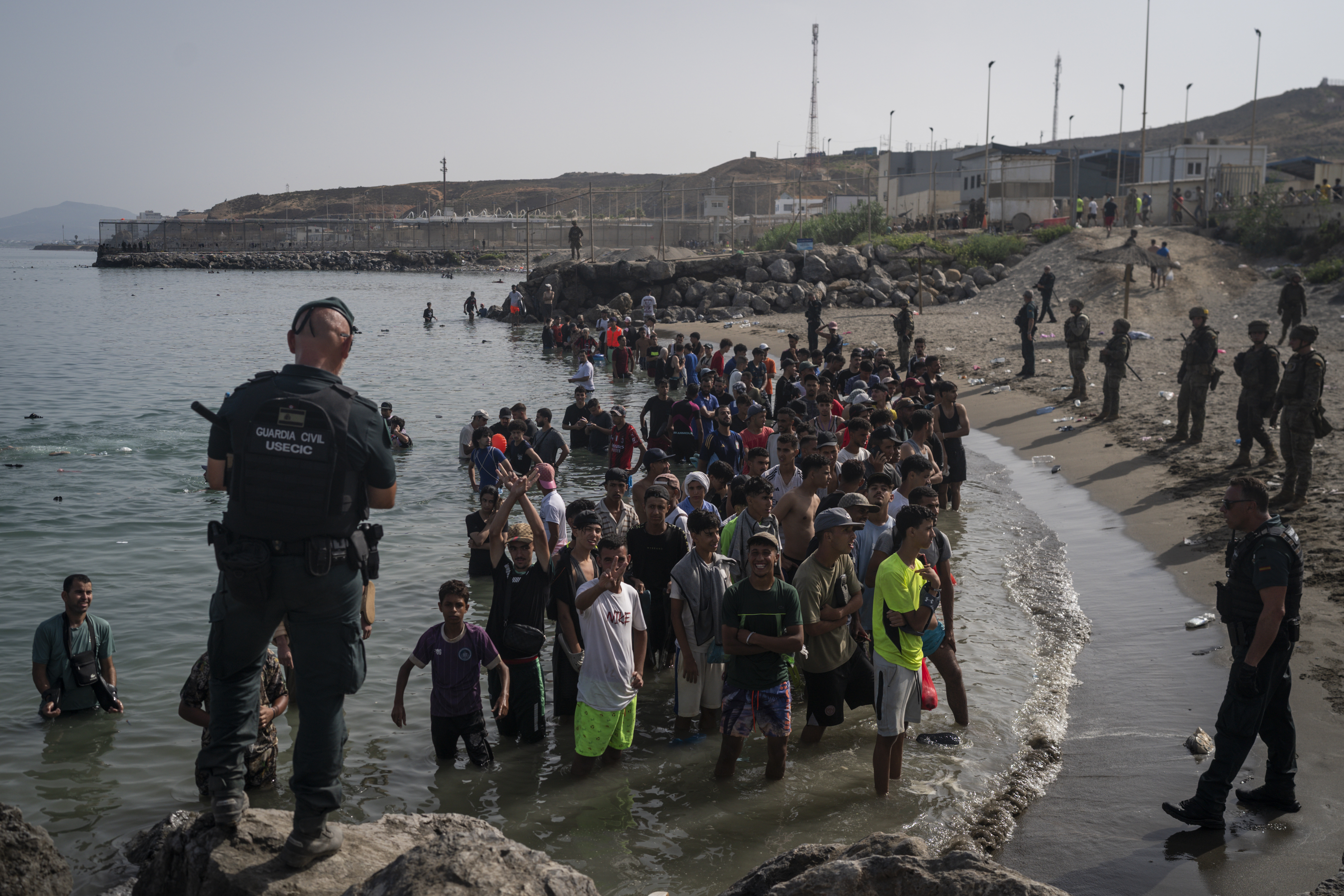 CEUTA, SPAIN - JULY 31: Members of the Guardia Civil USECIC guard migrants on Tarajal beach after crossing the border between Spain and Morocco on July 31, 2026 in Ceuta, Spain. Spain's interior ministry has said it's deploying 60 members of the armed forces and an additional 30 Guardia Civil officers to Ceuta after tens of thousands of migrants entered the Spanish enclave in North Africa on Thursday. A large number of people swam through the Tarajal breakwater to enter the Spanish territory from neighboring Morocco, while others scaled the border fence. Some estimates have said that around 49,000 migrants made the crossing, with the surge of arrivals overwhelming migrant reception centres, with many sleeping on the streets overnight. (Photo by Adri Salido/Getty Images)